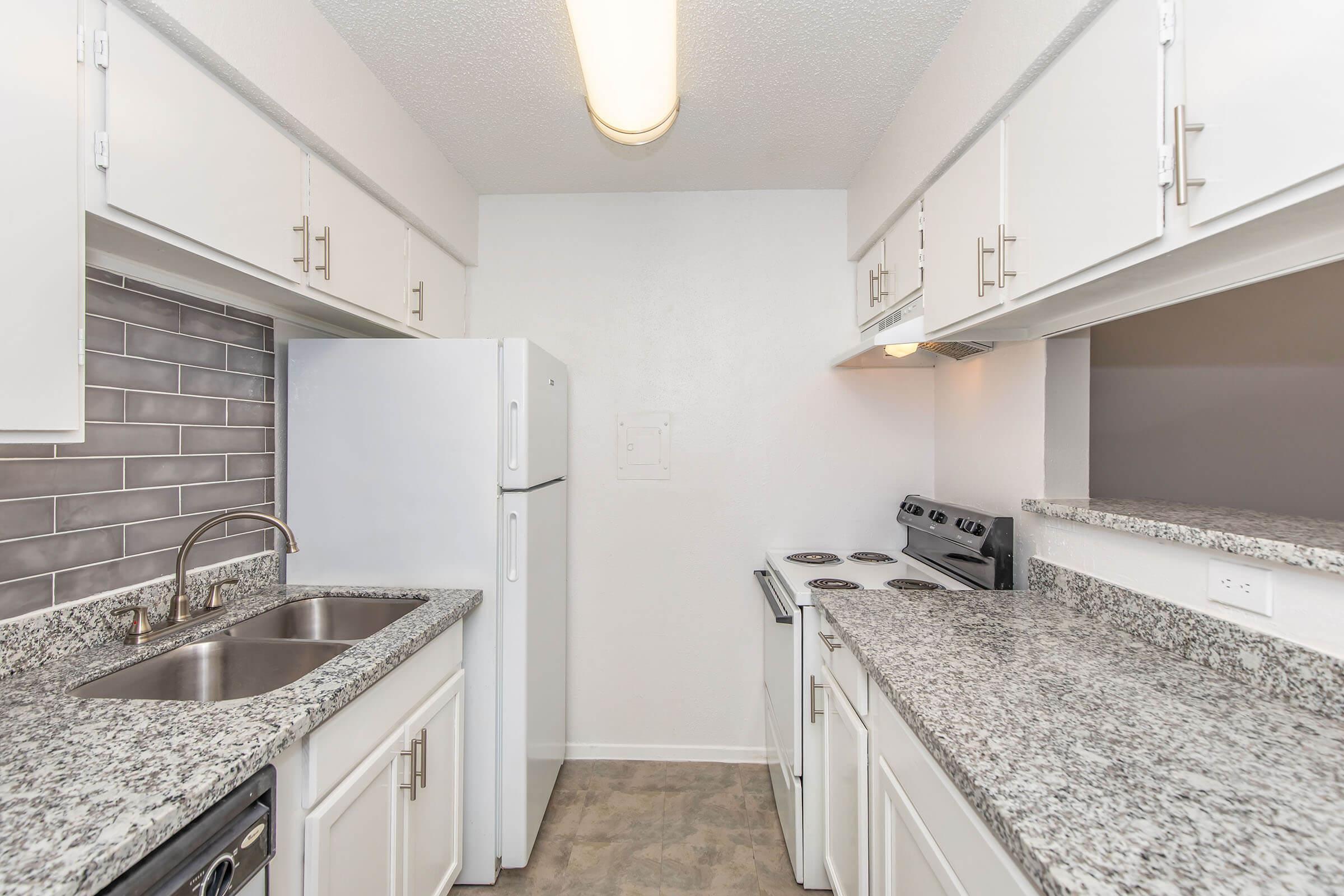 A modern kitchen featuring white cabinets, a stainless steel sink, a white refrigerator, an electric stove, and a backsplash with gray tiles. The countertops are made of granite, and there is a bright overhead light illuminating the space.