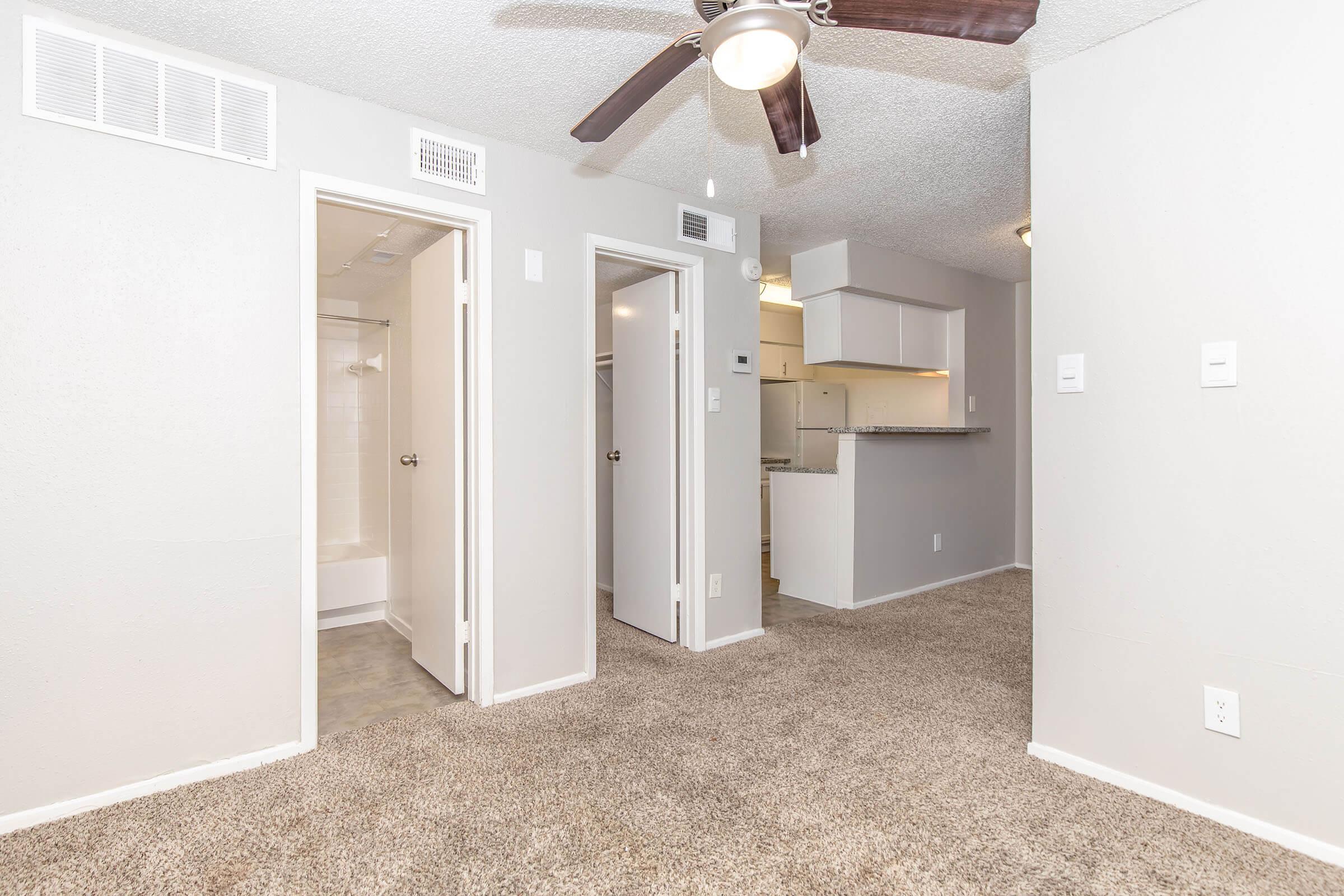 A well-lit interior of an apartment featuring a carpeted living area, two closed doors leading to rooms, and an open kitchen visible in the background. The walls are painted in a light color, and a ceiling fan is installed. The bathroom is partially visible to the left.