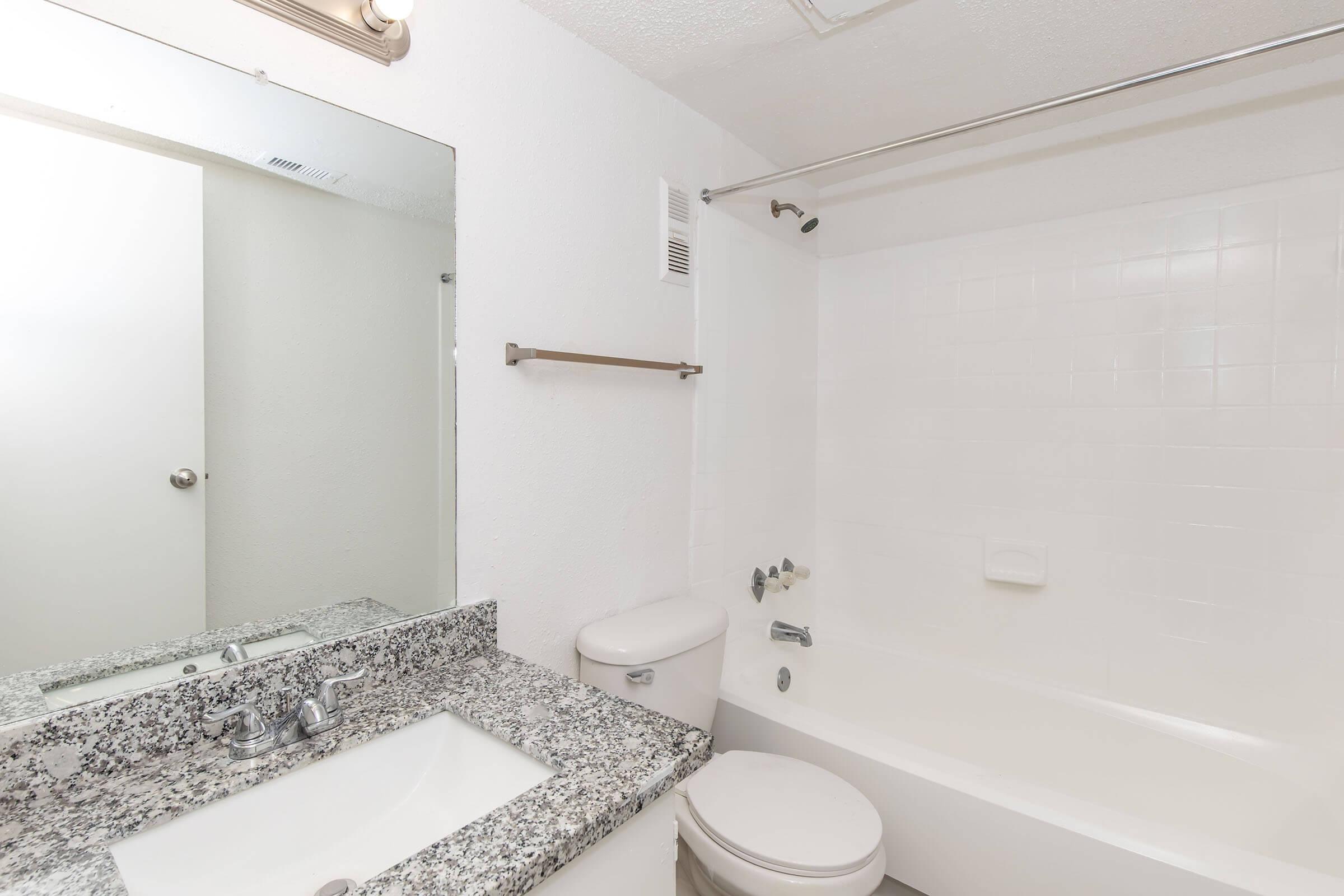 A clean, bright bathroom featuring a white bathtub, a modern toilet, and a sink with a granite countertop. A large mirror is mounted above the sink, and the walls are painted white. There's a towel bar next to the tub, and the room has a simple, minimalist design.