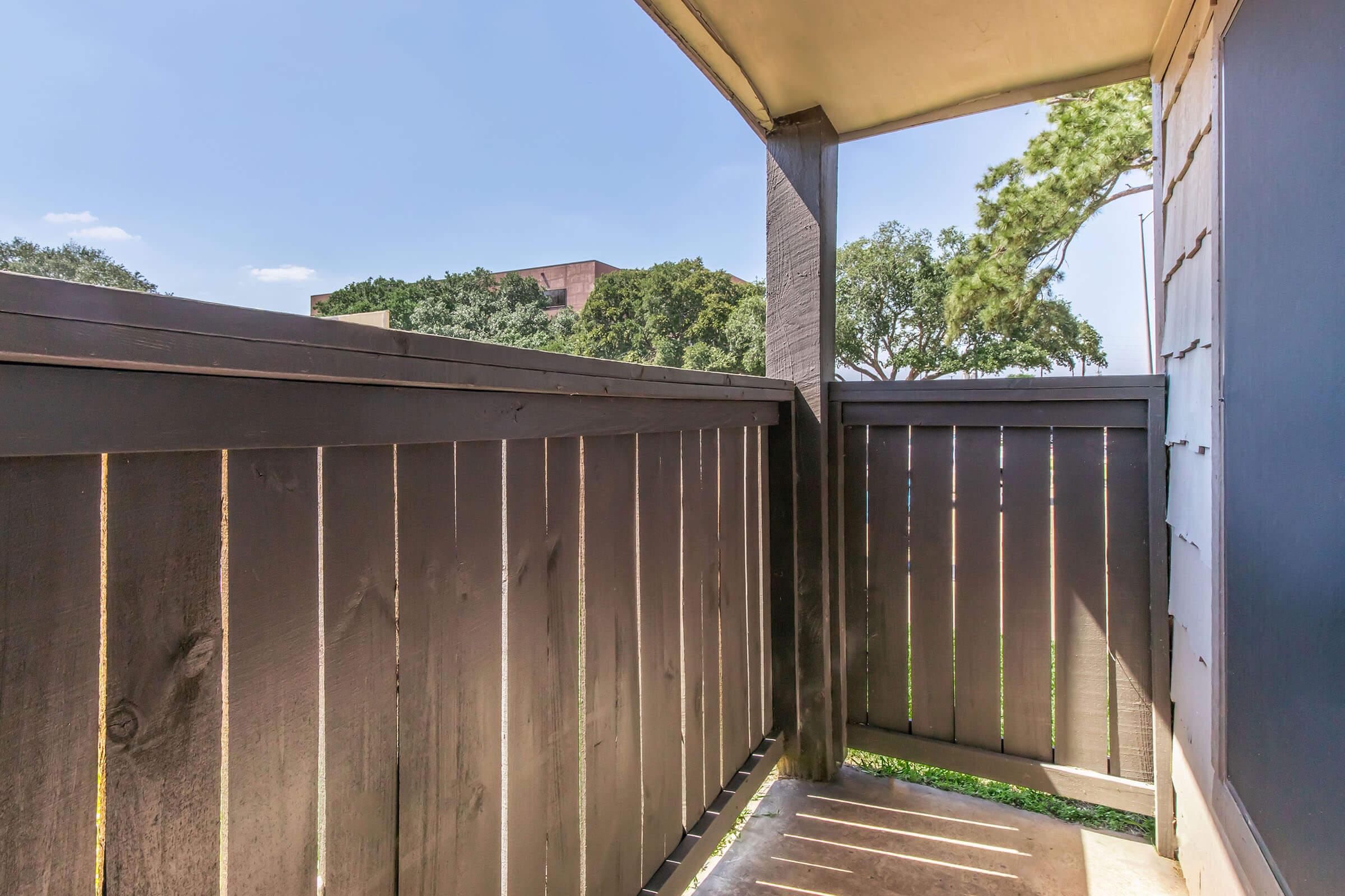 View of a balcony with dark wooden railing, looking out onto trees and a building in the distance. The sky is clear and blue, creating a bright atmosphere. The flooring is also wooden, and the scene suggests a quiet outdoor space for relaxation.