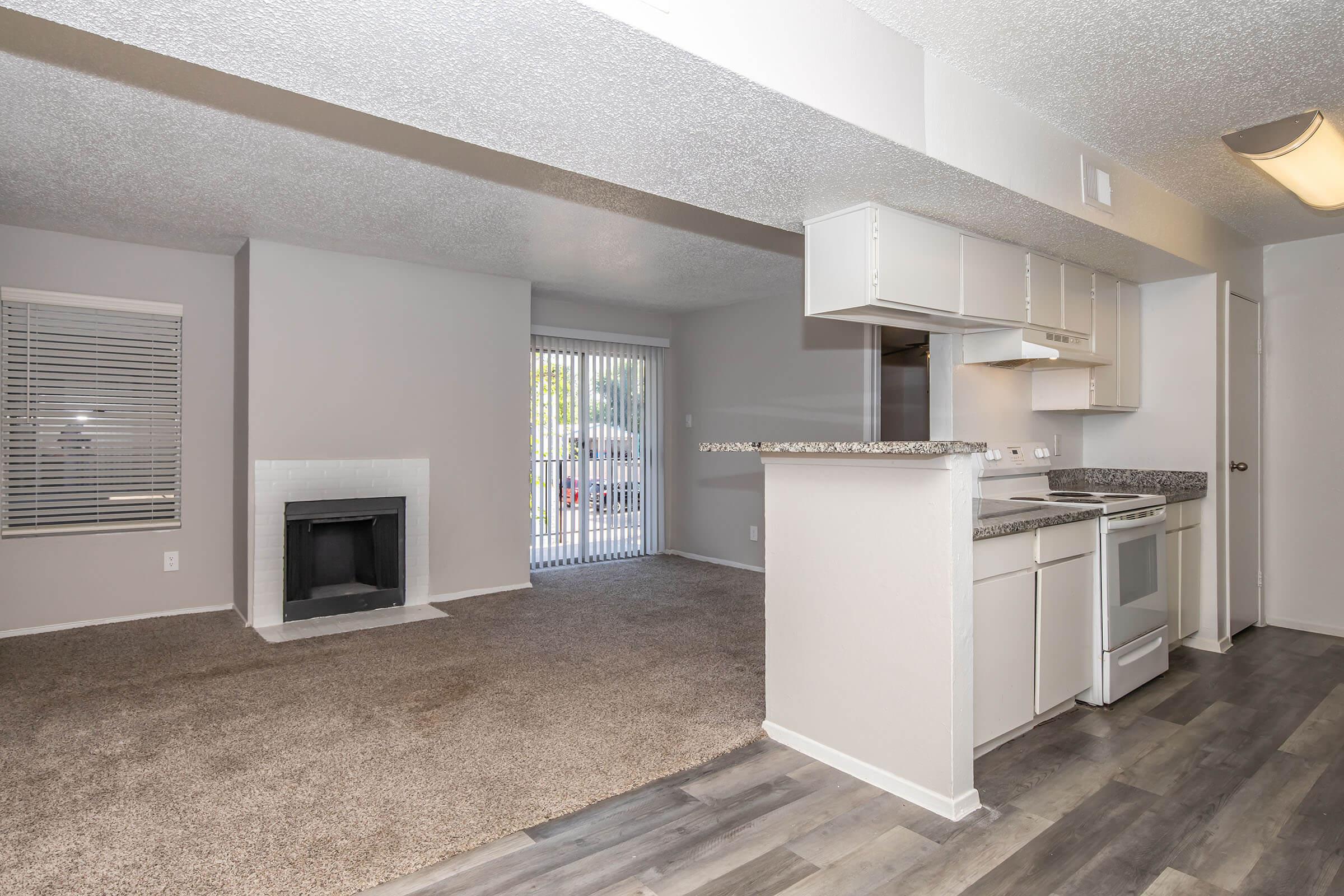 Interior of an apartment featuring an open layout. There is a kitchen with white cabinetry, a granite countertop, and appliances, including a stove and oven. Adjacent is a carpeted living area with a fireplace and a sliding glass door leading to a patio. Natural light fills the space, creating a welcoming atmosphere.