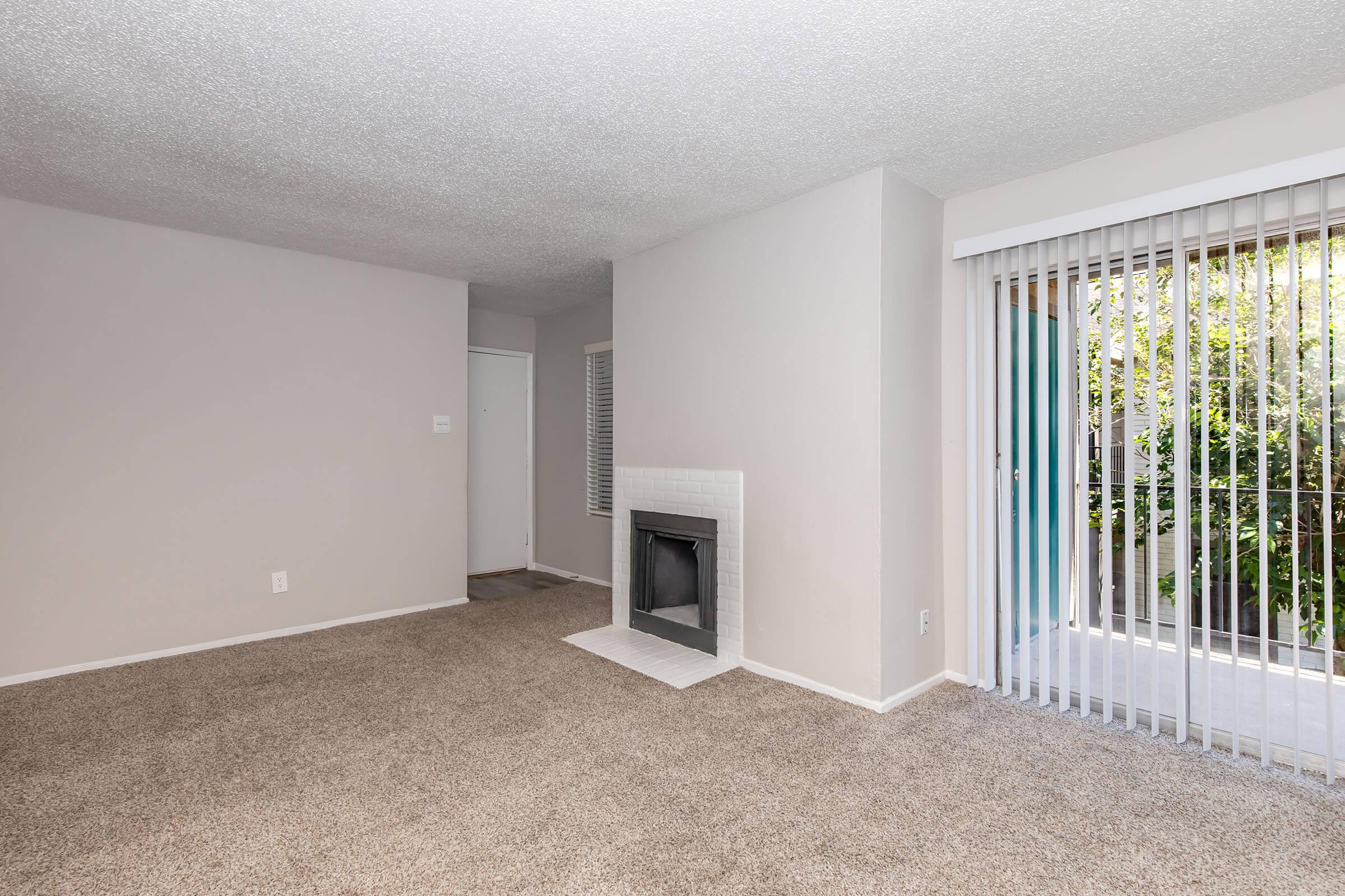 Spacious living area with light-colored walls and plush carpet. A large window with vertical blinds offers natural light, while a decorative fireplace adds a focal point. A doorway leads to another room, enhancing the open feel of the space.