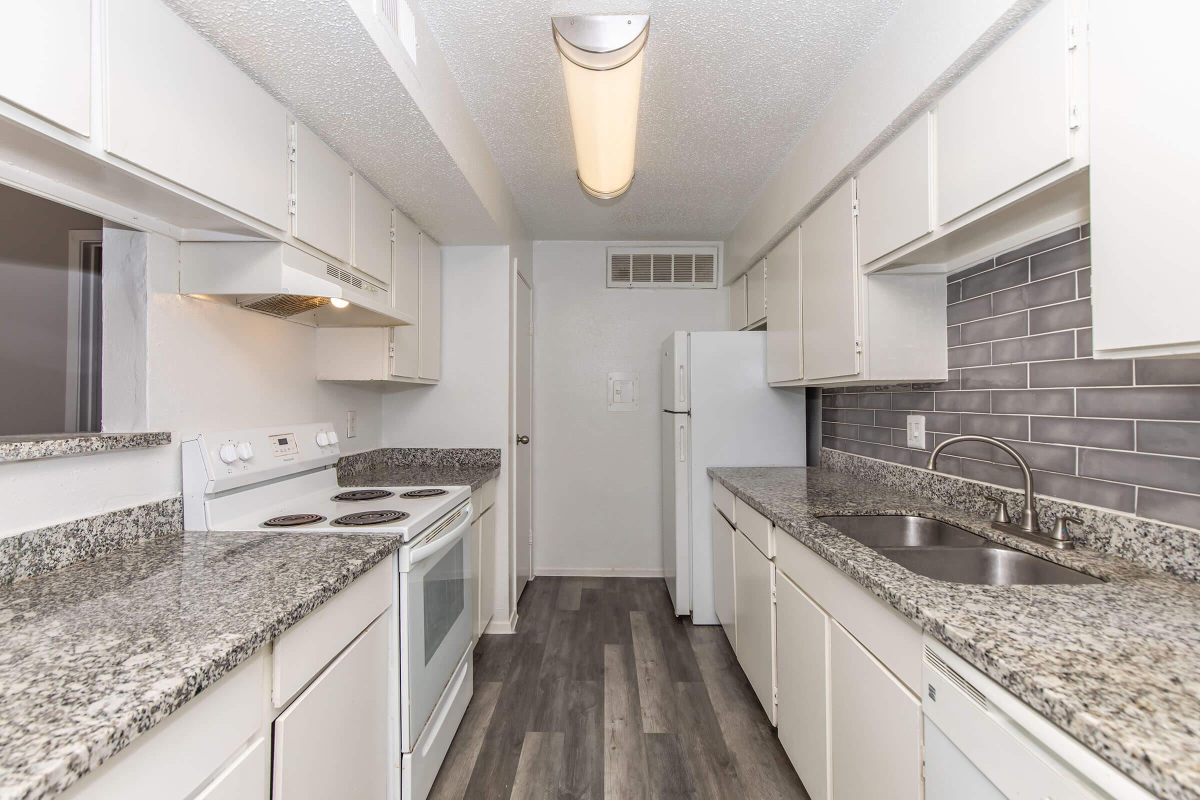 A modern kitchen with light-colored cabinets, granite countertops, and a tiled backsplash. It features a stove, oven, and refrigerator, with a sink and faucet. The flooring is a dark laminate, and there is overhead lighting providing ample illumination. The layout is open and functional.