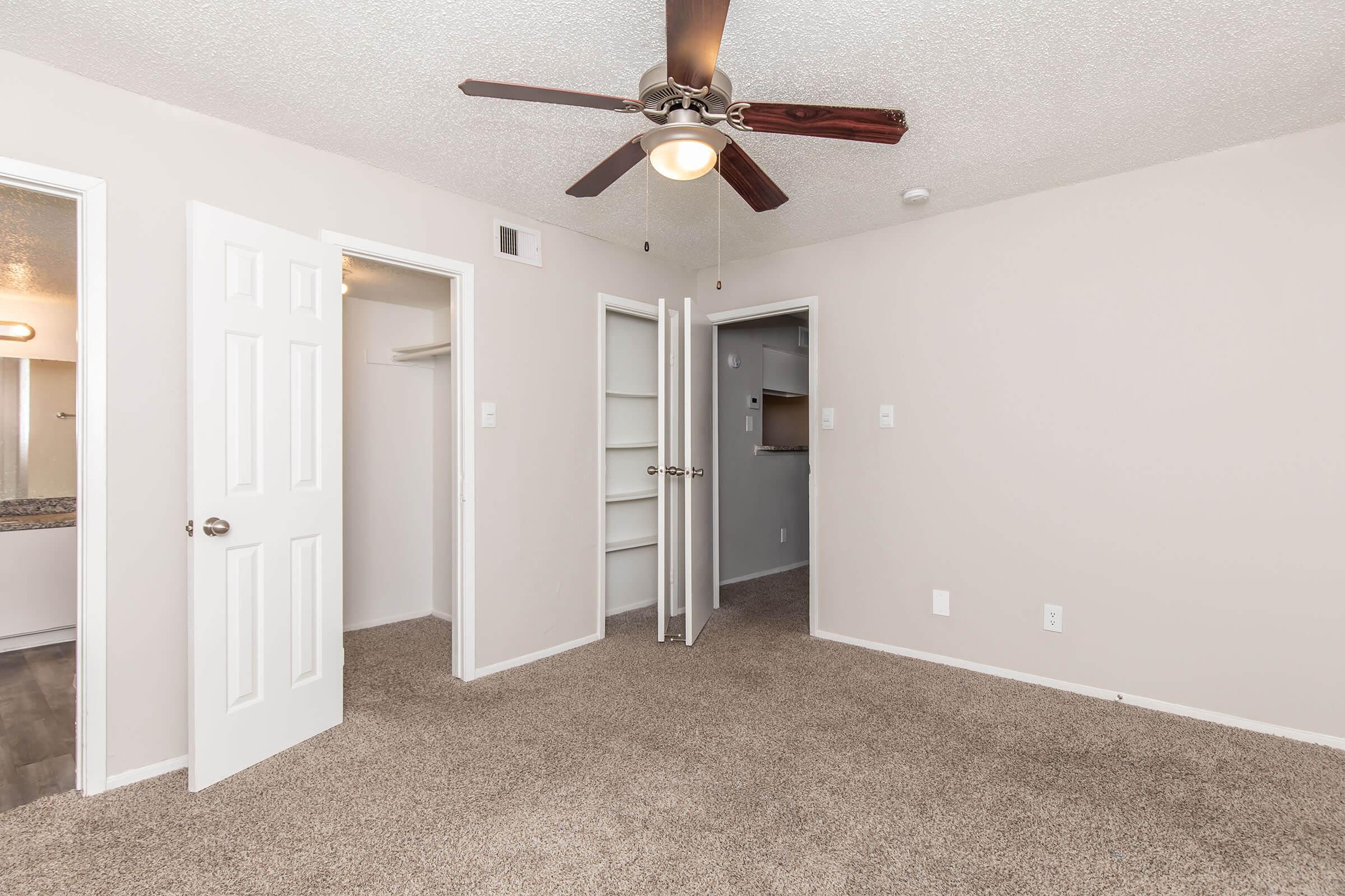 Interior view of a small, neutral-colored bedroom featuring carpeted floors, a ceiling fan with light, and two doors leading to closets. One closet is open, revealing shelves. The walls are plain, and there's a slight indication of light coming from an adjoining room.