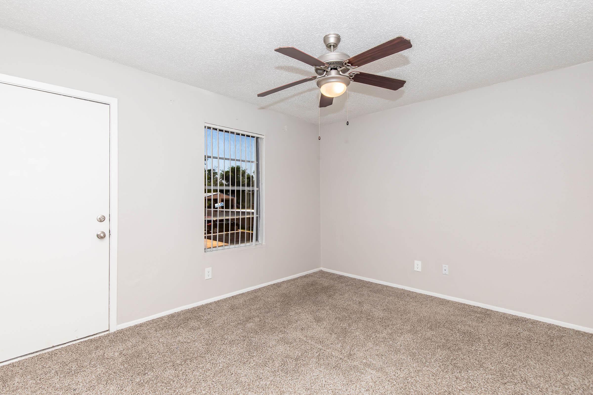 A small, empty room featuring light beige carpeting, a ceiling fan with a light fixture, and a window with metal bars. The walls are painted a light color, and there is a closed white door. The space is bright and uncluttered, suitable for various uses like a bedroom or office.