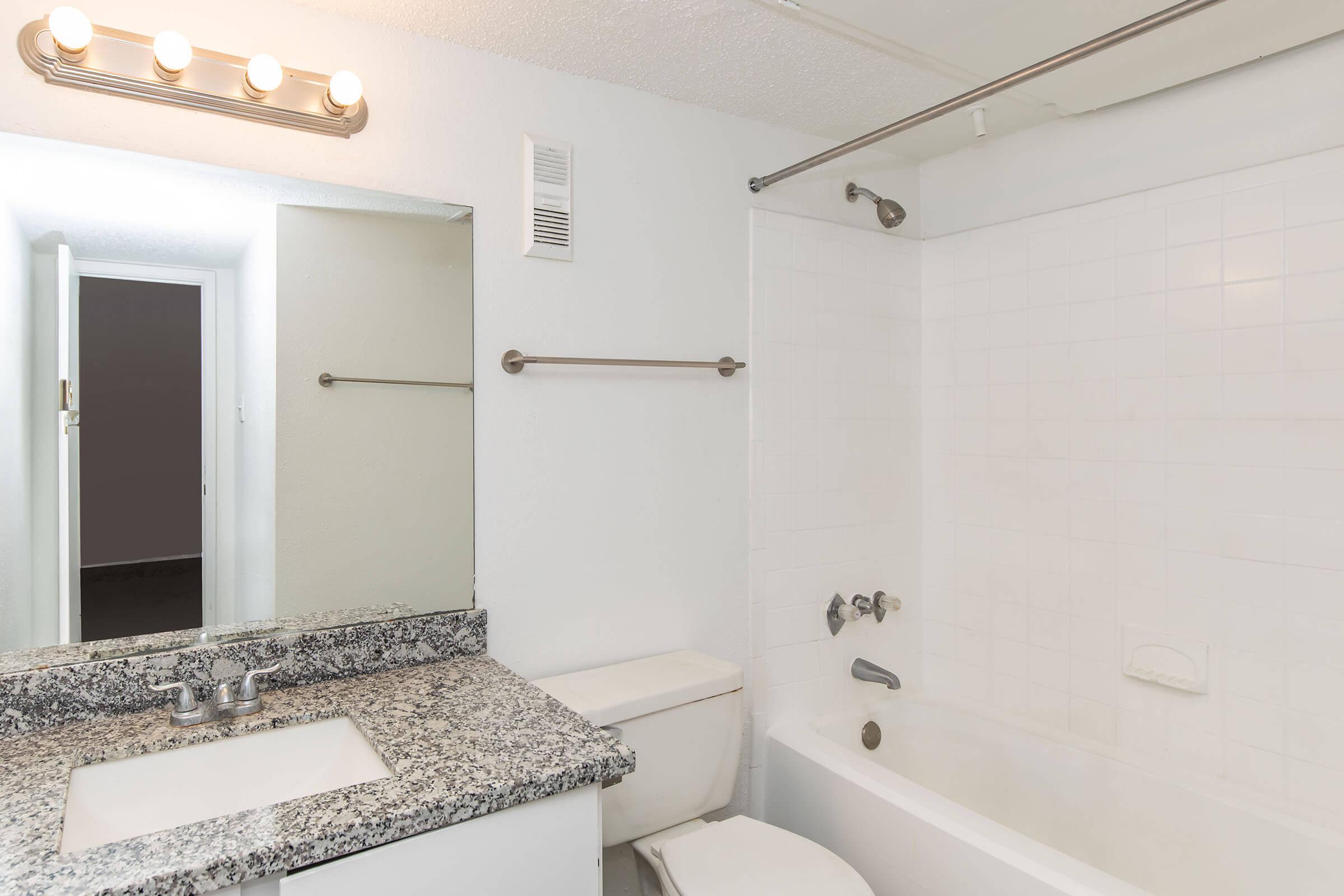 A modern bathroom featuring a white tiled shower and bathtub, a granite countertop with a sink, a mirror above the sink, and a towel bar. The walls are painted white, and there is bright overhead lighting. A door is partially visible, leading to another room.