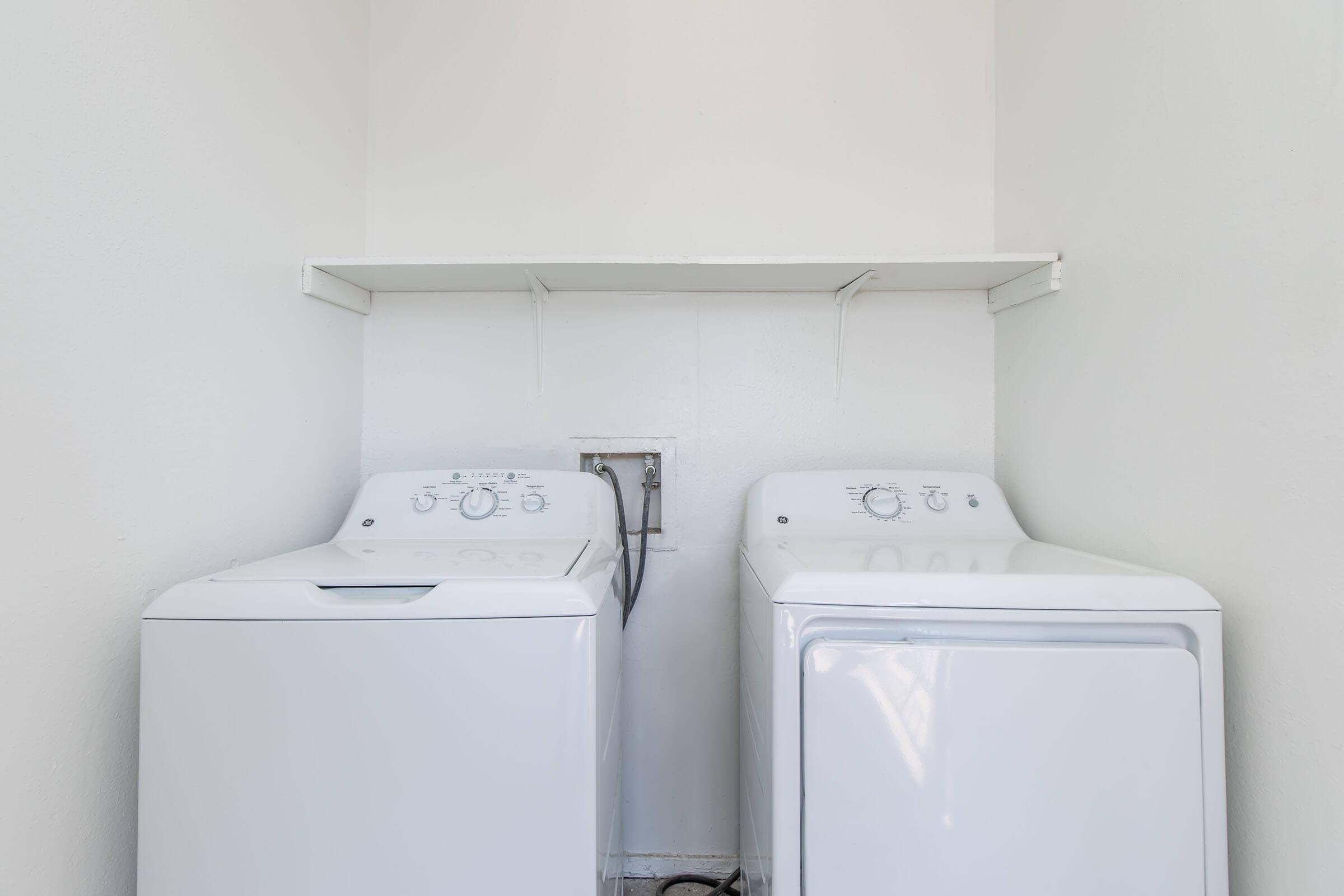 A bright laundry area featuring a white washing machine and a white dryer side by side, with a shelf above them. The walls are plain and light-colored, creating a clean and simple aesthetic. The space is uncluttered, emphasizing the appliances.