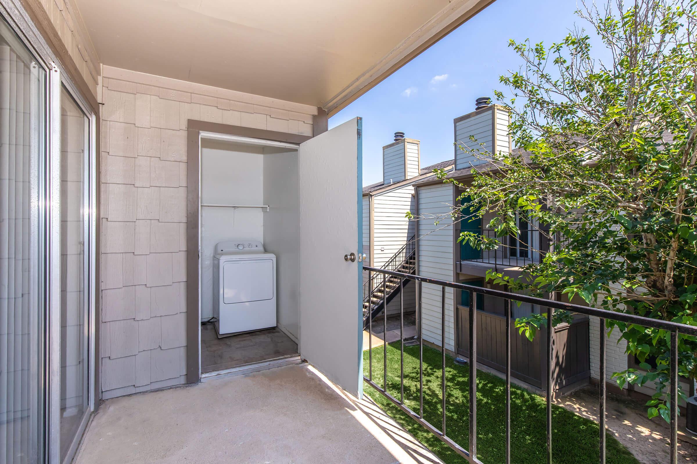 A balcony view featuring an open door leading to a laundry area with a washing machine. The scene includes greenery and adjacent apartment buildings, under a clear blue sky.