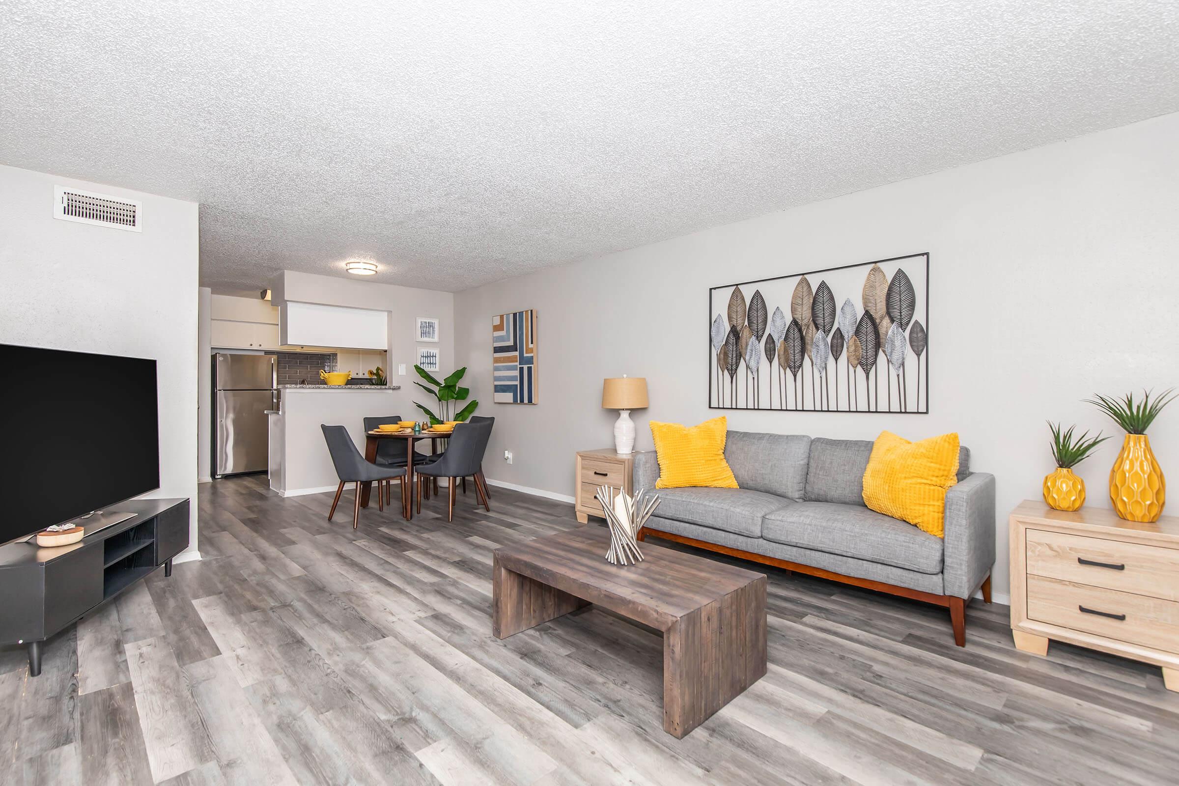 A modern living room featuring a gray sofa with yellow cushions, a wooden coffee table, and a television. In the background, there's a dining area with a table and chairs, a plant, and decorative vases. The space has a light, airy feel with light wood flooring and neutral wall colors.