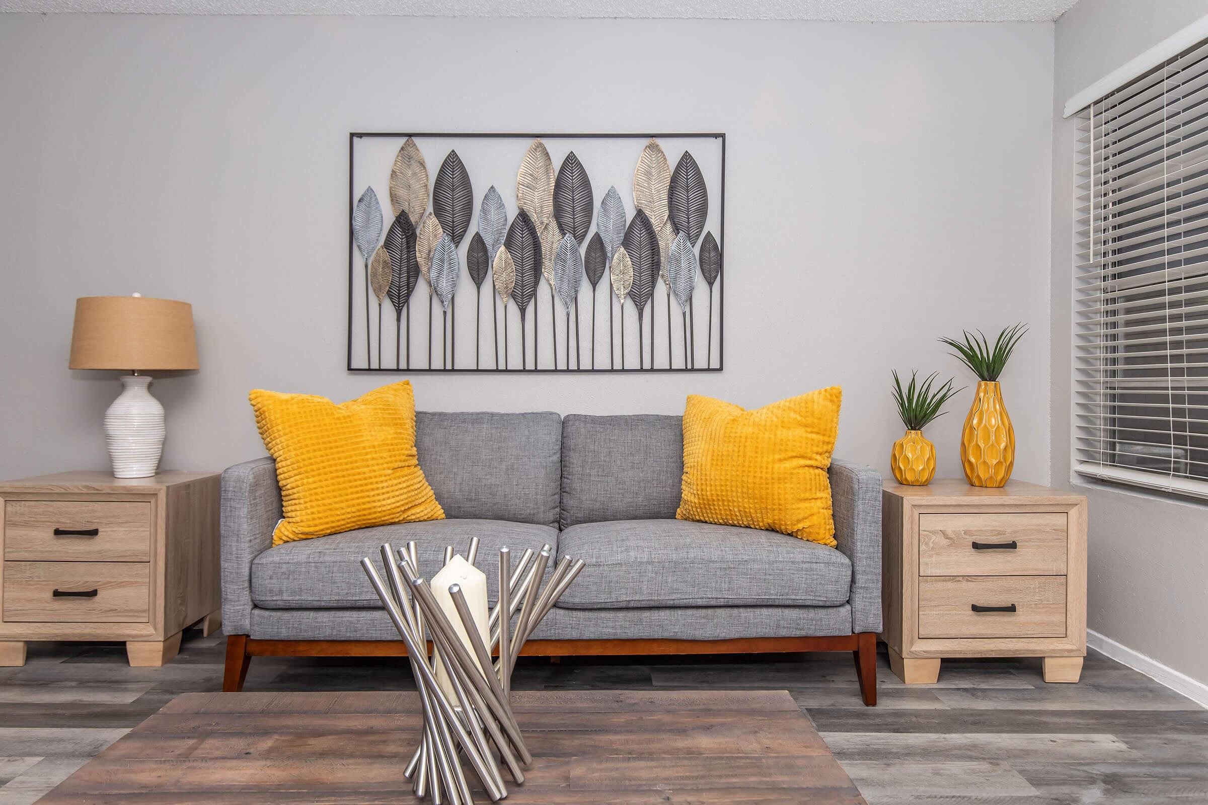 A modern living room featuring a gray couch with two yellow cushions, a wooden coffee table with metallic decor, two light wooden side tables, a decorative yellow vase, and a wall art piece displaying stylized leaves. The room has neutral walls and a large window with blinds.