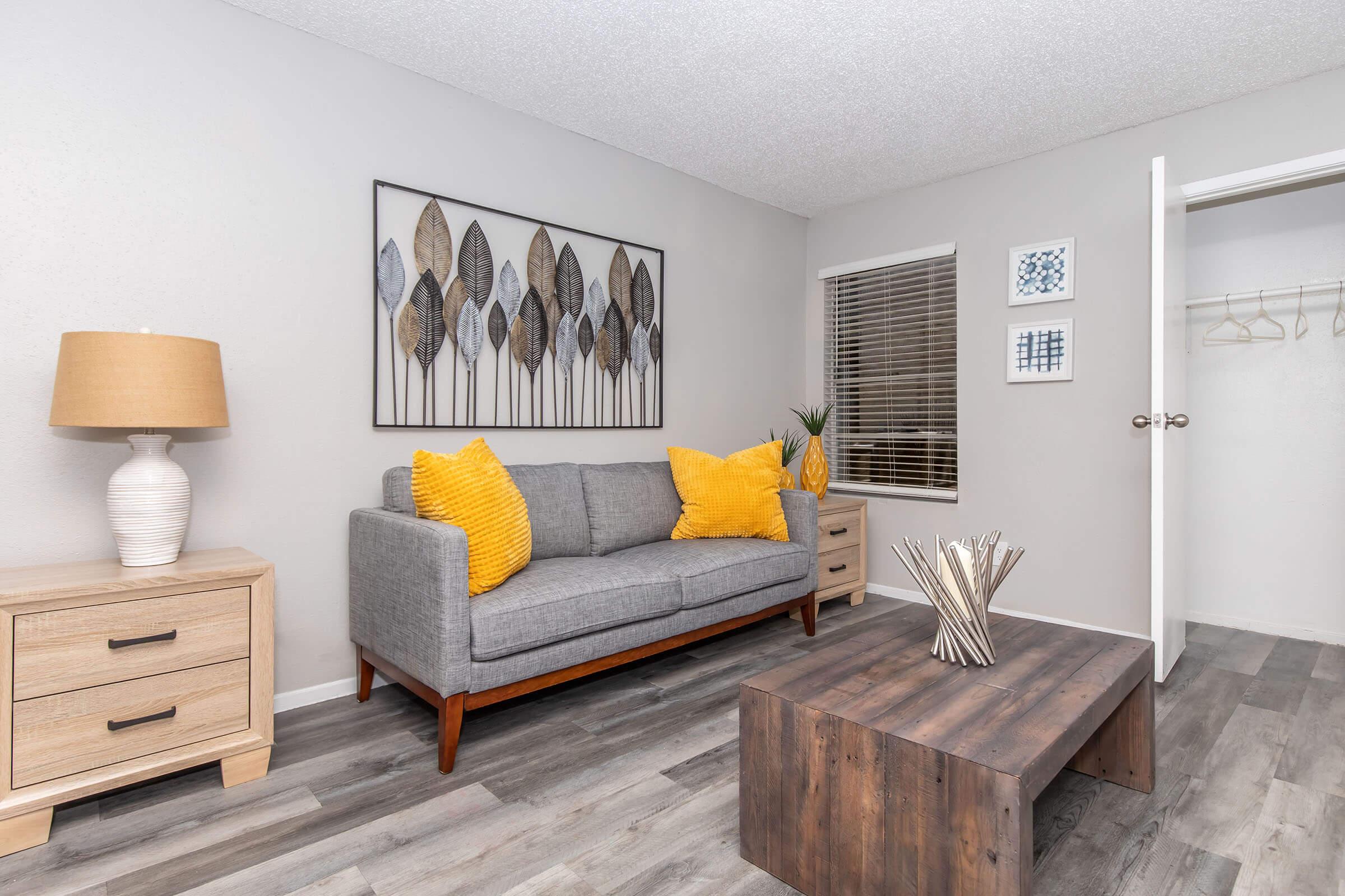 A modern living room featuring a gray sofa with yellow cushions, a wooden coffee table, and a lamp on a side table. The walls are light gray with decorative art showcasing leaf designs. A window with blinds allows natural light, and a closet door is partially open, revealing hangers inside.