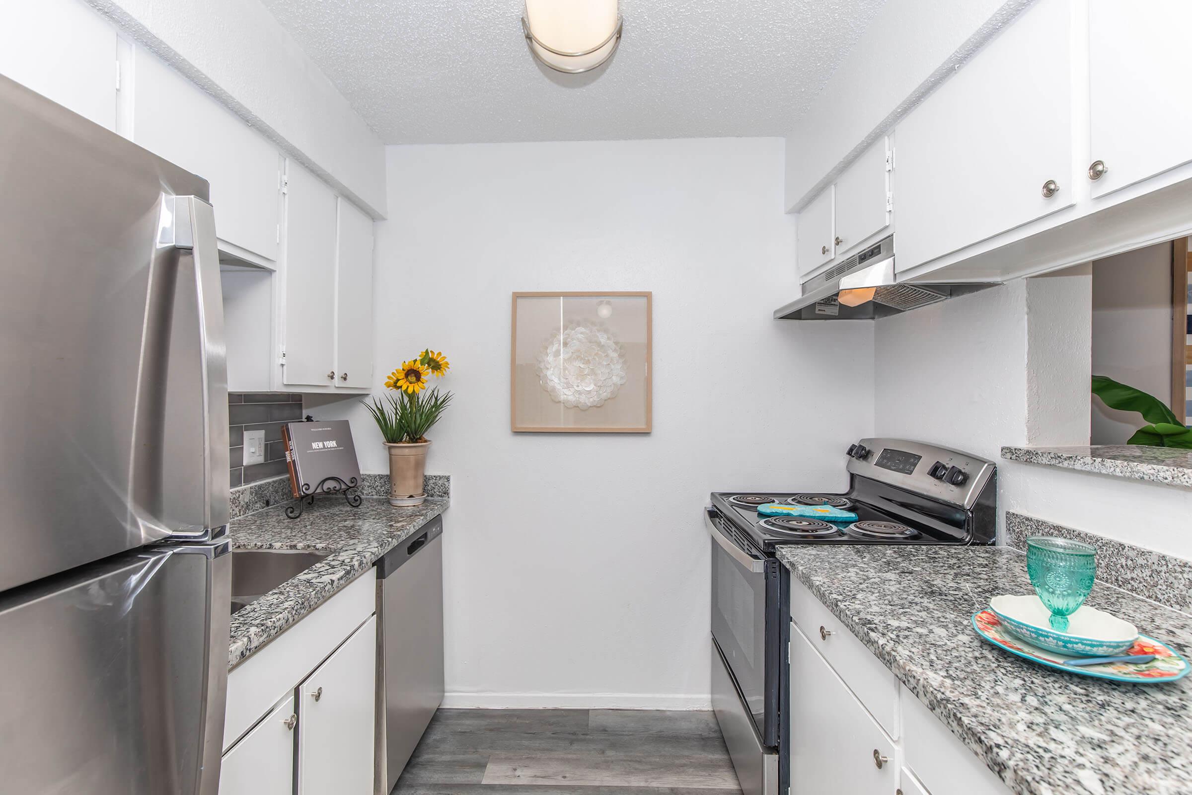 A modern kitchen featuring stainless steel appliances, including a refrigerator and stove. The countertops are made of granite, and there is a decorative wall art piece above. Sunflowers in a vase add a touch of color, while a dining set with turquoise plates is arranged on the counter. The overall design is clean and bright.