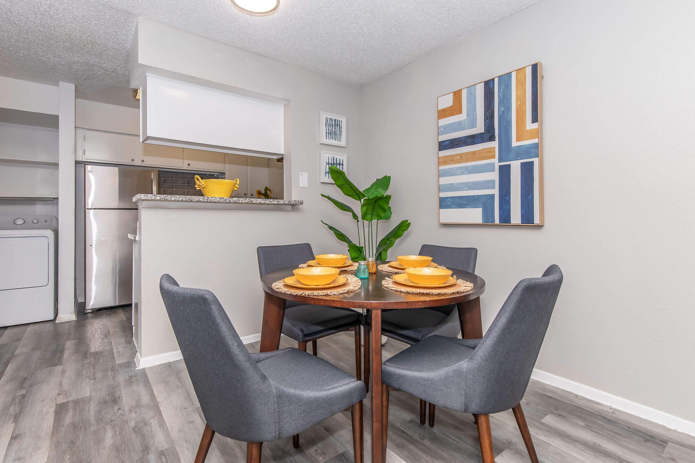 A modern dining area featuring a round wooden table set with four yellow bowls and decorative coasters. Surrounding the table are four gray upholstered chairs. In the background, there is a kitchen area visible with appliances, and a piece of abstract wall art hangs beside a green plant on the table.