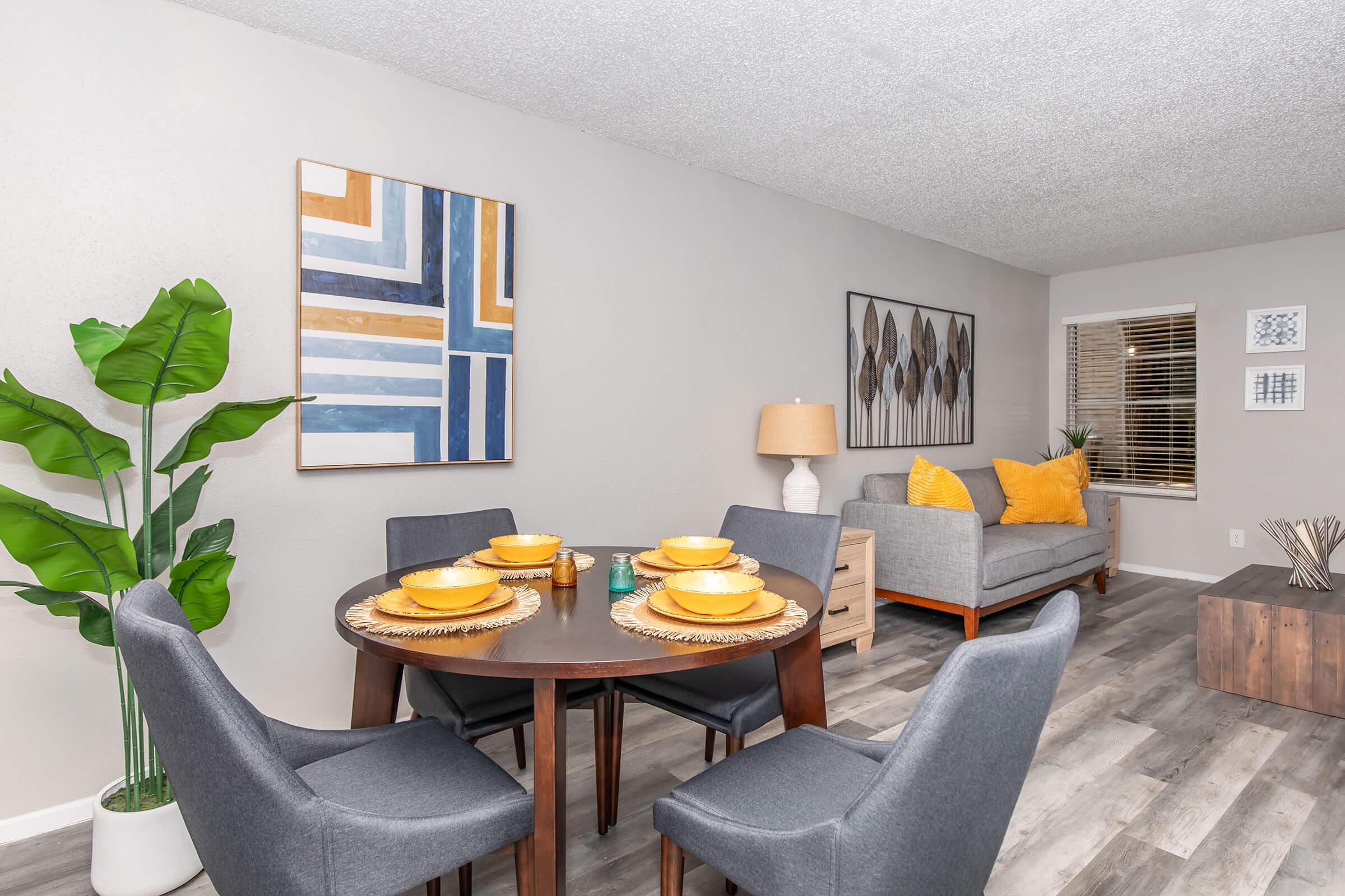 A cozy dining area featuring a round table set with yellow dishes and woven placemats. Four gray chairs surround the table. In the background, a gray sofa with yellow throw pillows is seen next to a wooden side table. Decorative wall art and a potted plant add to the inviting ambiance of the space.