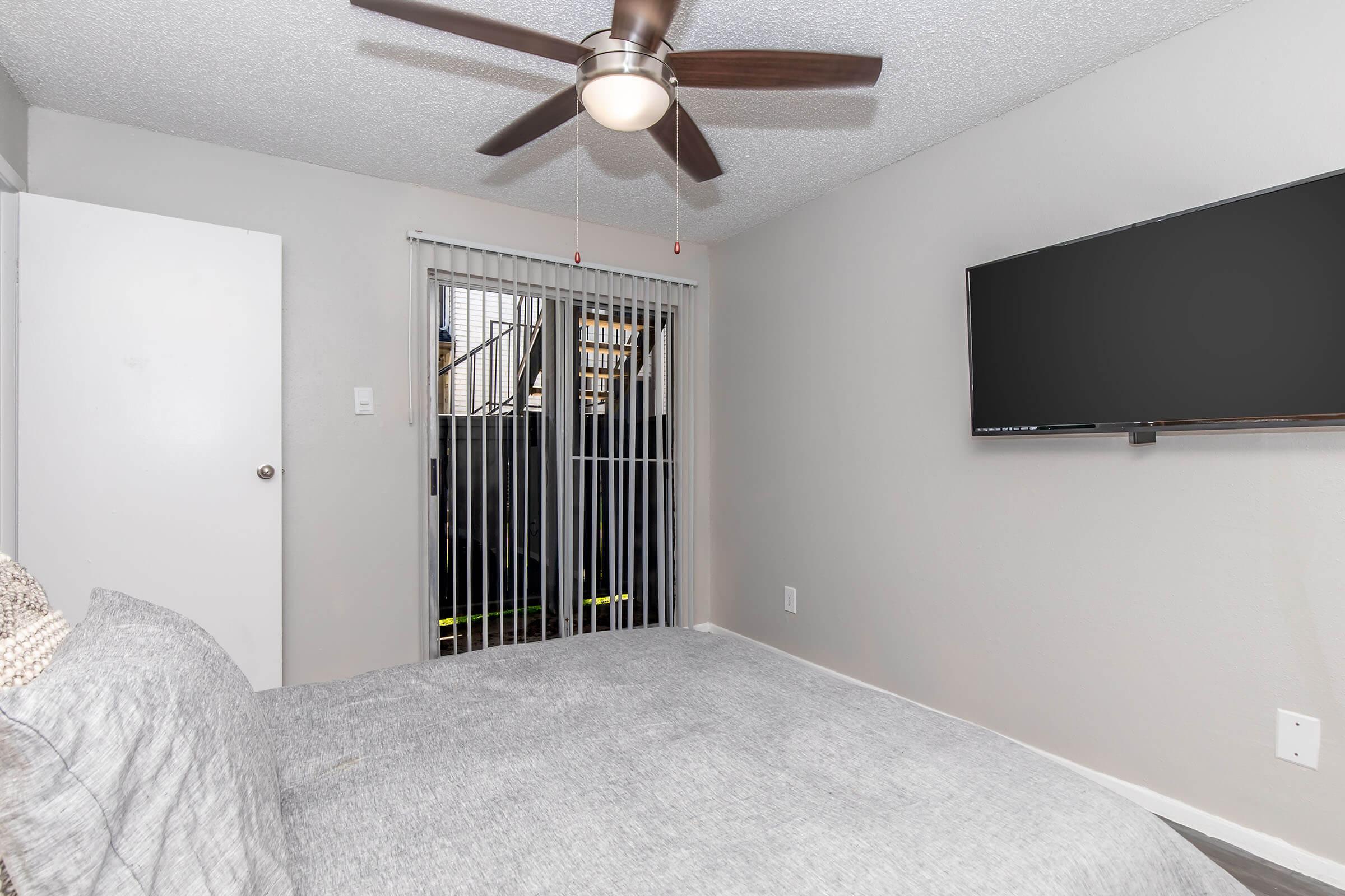 A cozy bedroom featuring a gray bed and a ceiling fan. There's a wall-mounted flat-screen TV and sliding glass doors with vertical blinds leading to an outdoor area. The walls are painted in a light color, creating a bright and airy atmosphere. A white door is visible on the left side of the room.