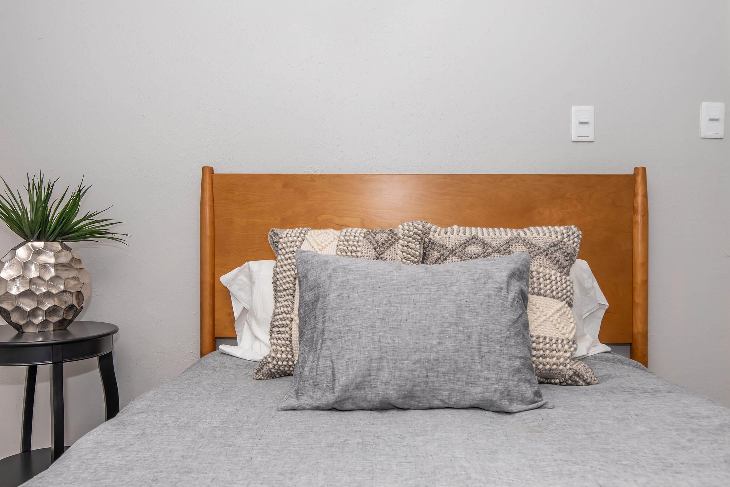 A neatly made bed with a wooden headboard, featuring various decorative pillows in neutral tones. A small side table with a decorative plant and a textured, silver lamp is positioned beside the bed against a light gray wall.