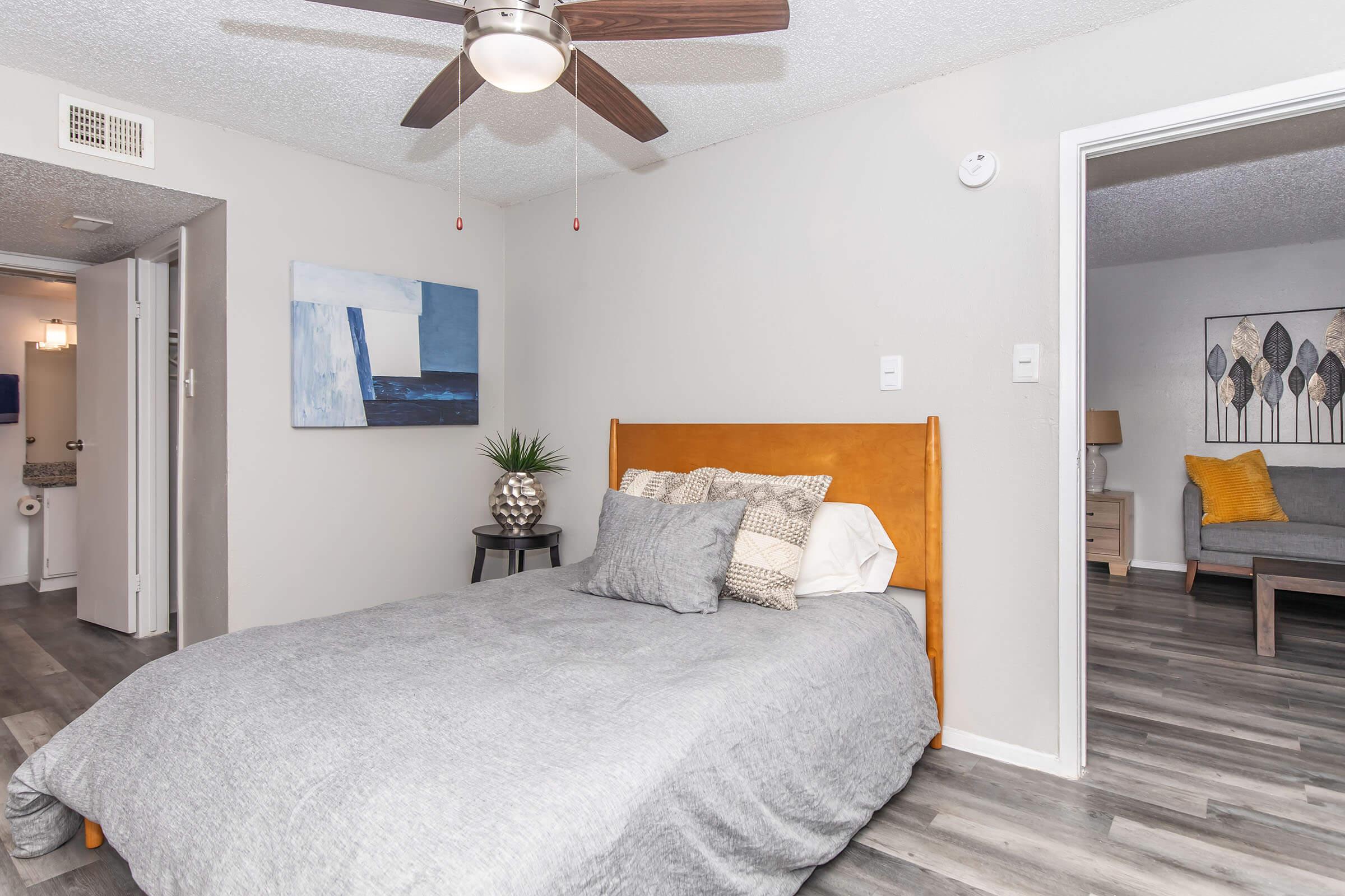 A cozy bedroom featuring a wooden bed with gray bedding and decorative pillows. A small nightstand holds a potted plant. The room has a ceiling fan and light gray walls. An open doorway leads to another room with a gray sofa and modern artwork on the walls. Natural light is present, creating a welcoming atmosphere.