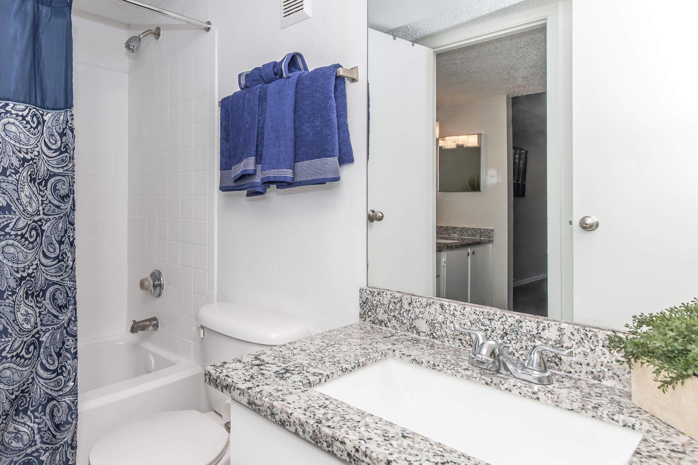 A modern bathroom featuring a white bathtub with a blue patterned shower curtain, a granite countertop with a sink, and neatly arranged blue towels hanging on a towel rack. A mirror is mounted above the sink, reflecting the light and bright decor. A small plant adds a touch of greenery.