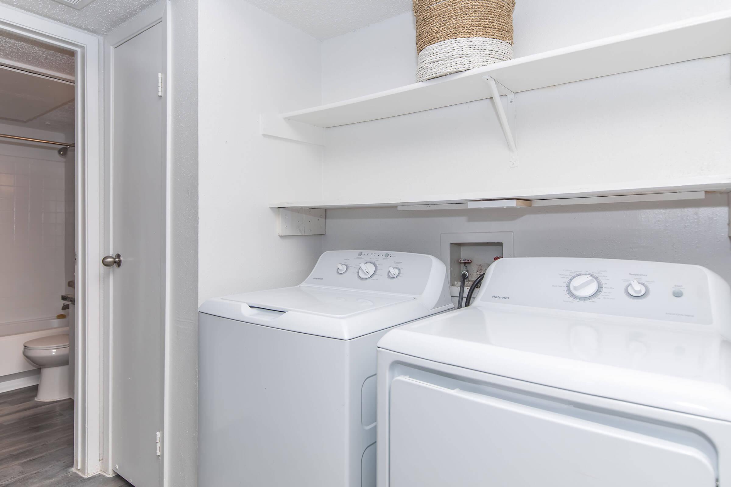 A laundry room featuring a washing machine and dryer side by side on a shelf, with white walls and a light gray floor. There is a door leading to another room in the background, and a woven basket placed on the shelf above the appliances.