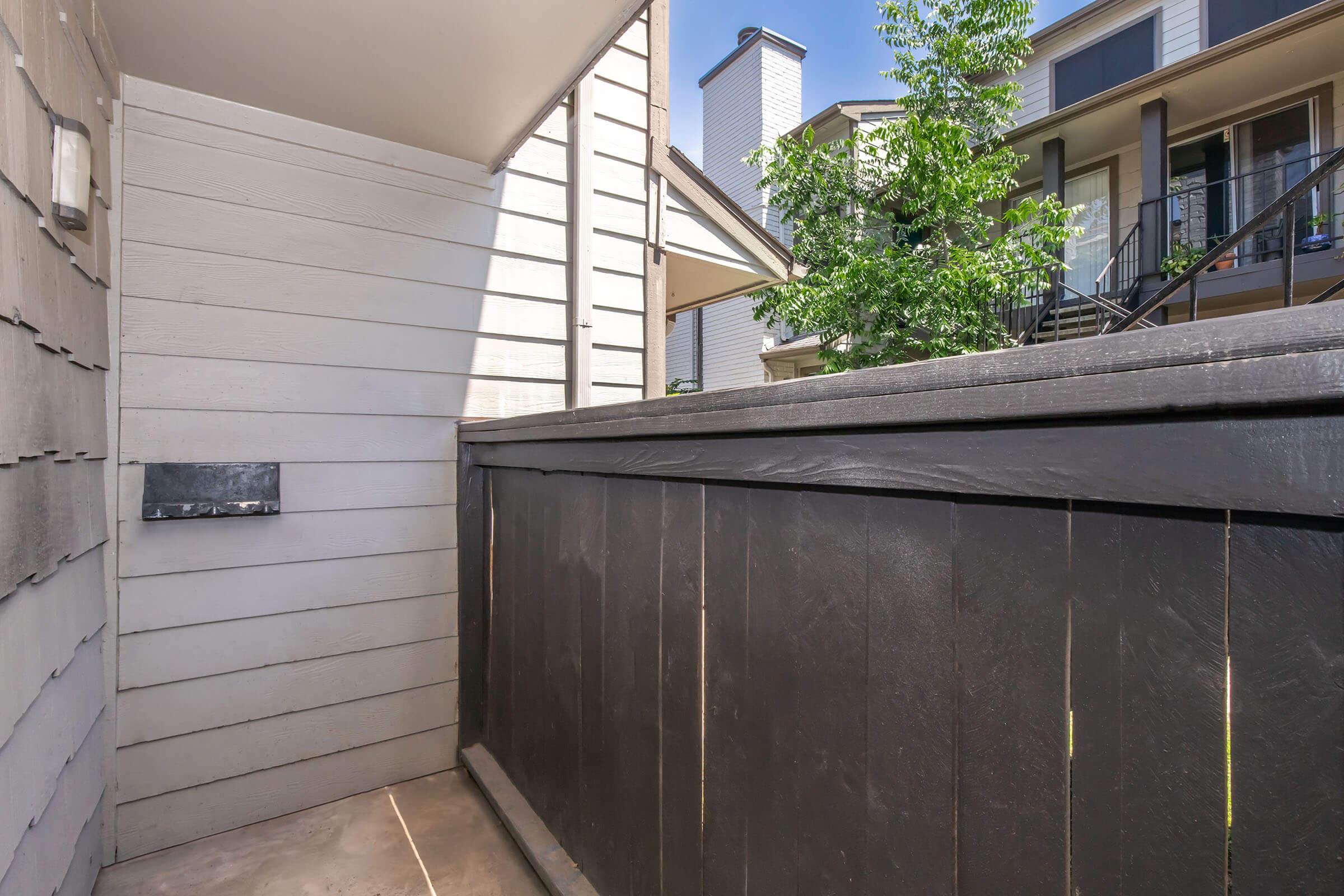 A corner view of an outdoor area featuring a wooden fence and light-colored siding. A mailbox is mounted on the wall, and greenery from nearby trees is visible in the background, along with the architecture of adjacent buildings.