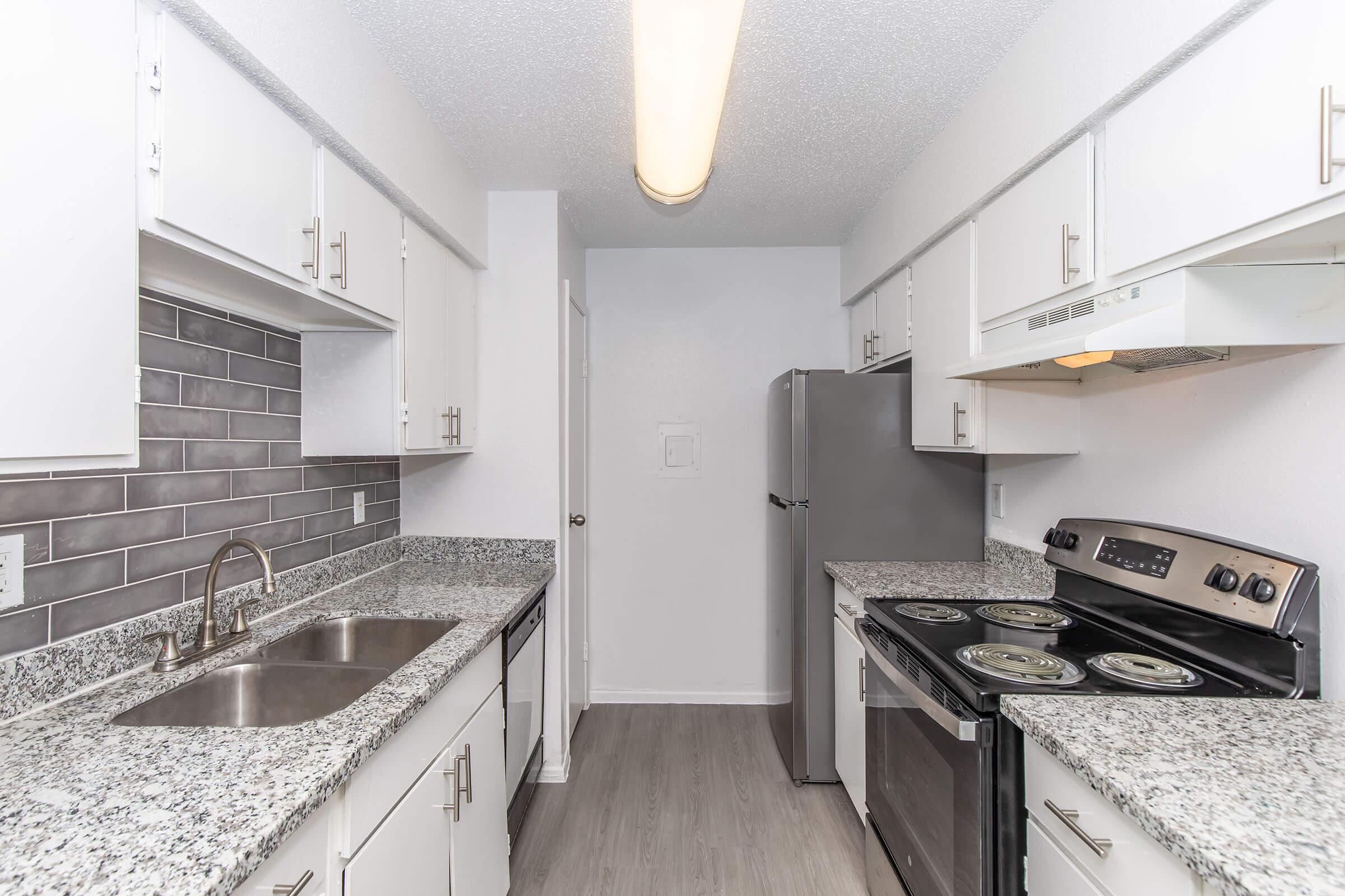 Modern kitchen with gray and white color scheme. Features a granite countertop, stainless steel appliances including a refrigerator and stove, dual sink with a faucet, and a tiled backsplash. The cabinets are white, and the flooring is a light wood finish. Bright overhead lighting enhances the space.