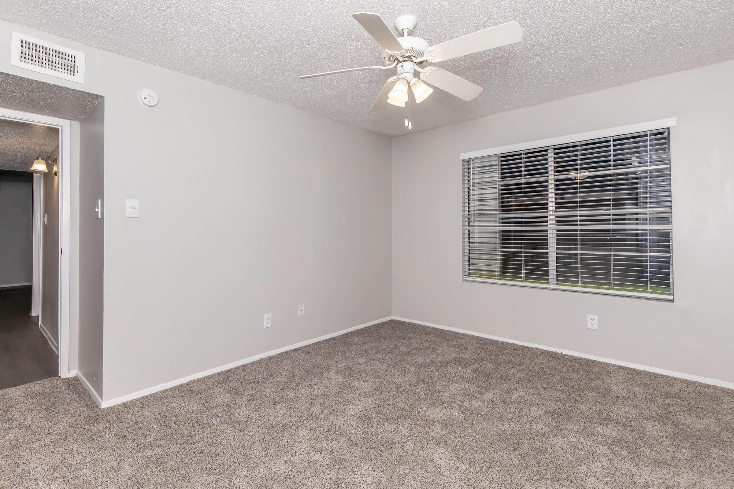 Empty room with light gray walls and carpet. A ceiling fan with three blades hangs in the center, and a large window with blinds allows natural light to enter. A doorway is visible on the left side, leading to another space. The room appears clean and well-maintained, suitable for various uses.