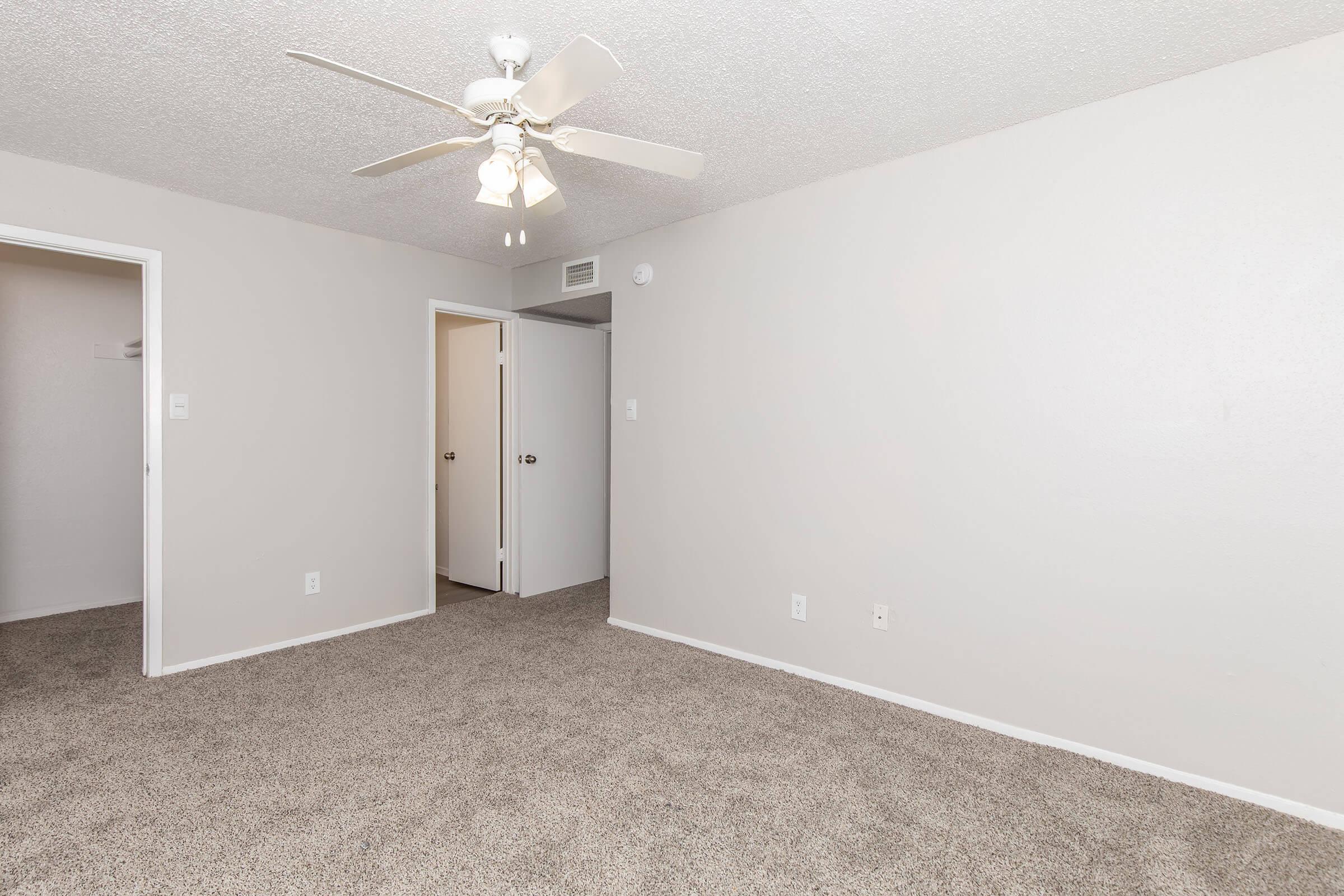 A spacious room featuring light gray walls and plush carpeting. A ceiling fan with white blades hangs from the ceiling. Doorways lead to adjacent rooms, with a simple design and neutral color scheme, creating a clean and inviting atmosphere. Natural light may brighten the space.