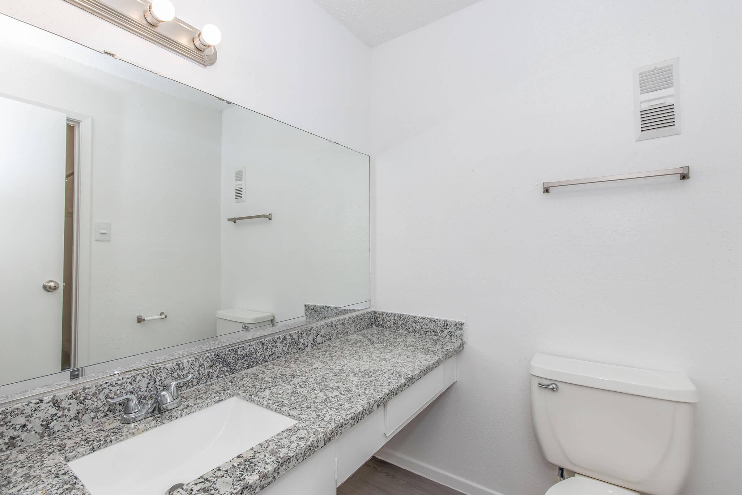 A clean and modern bathroom featuring a granite countertop with a sink, a large mirror, a toilet, and a towel bar. The walls are painted white, and there is a wall vent. Natural light is complemented by overhead lighting. The floor appears to be a light wood finish.