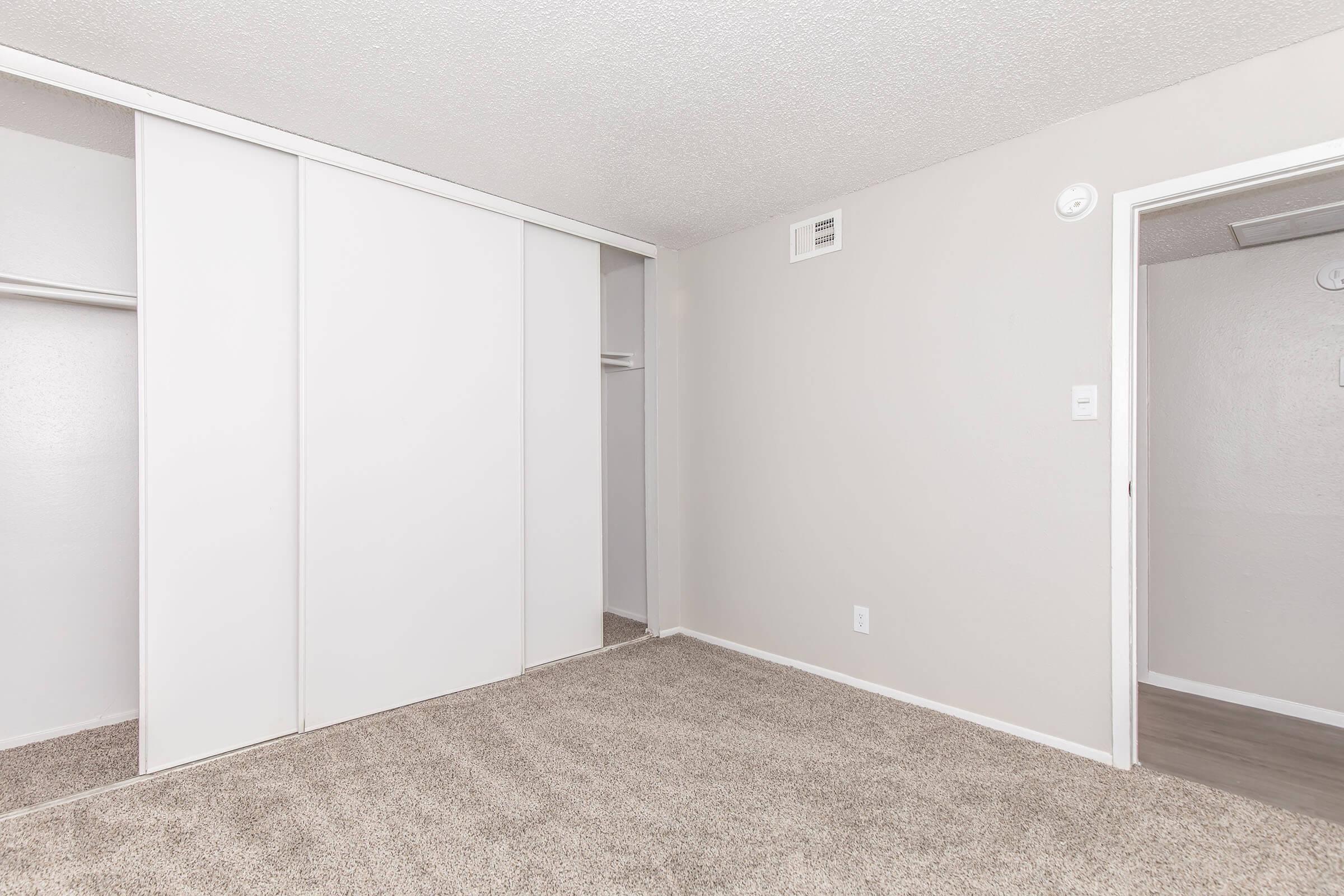 A spacious, empty room with light gray walls and carpet. There is a closet with sliding white doors on the left, and a doorway leading to another room on the right. The overall ambiance is neutral and clear of furniture, creating a bright and inviting space.