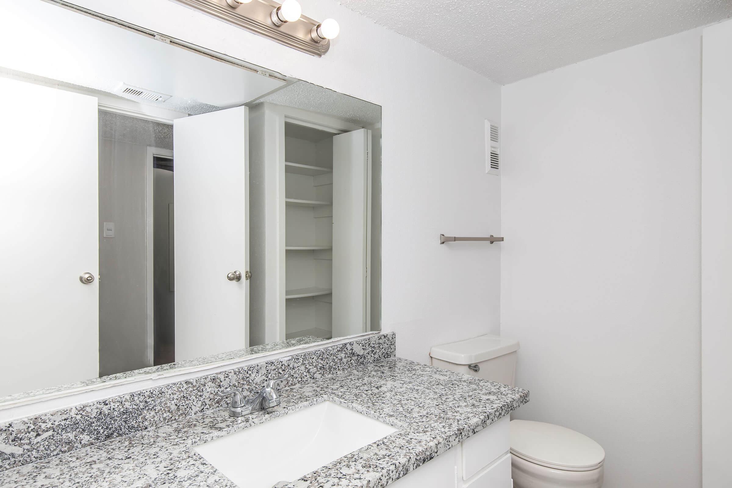 A clean, modern bathroom featuring a granite countertop with a sink, a large mirror above, and a toilet. The walls are painted white, and there are two sliding doors revealing a storage closet. A towel bar is mounted on the wall, and natural light illuminates the space.