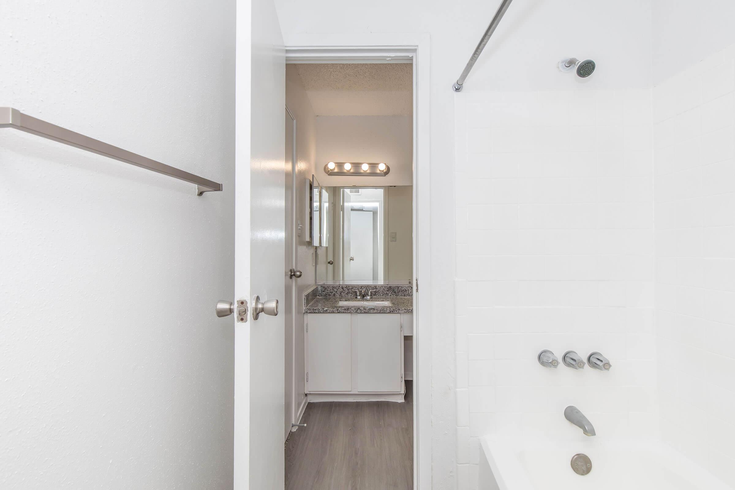 A clean, well-lit bathroom with a partially open door leading to a vanity area and a shower tub. The bathroom features white walls, a mirror with lights above the sink, and a towel rack. The floor has a light-colored finish, creating an airy and modern feel.