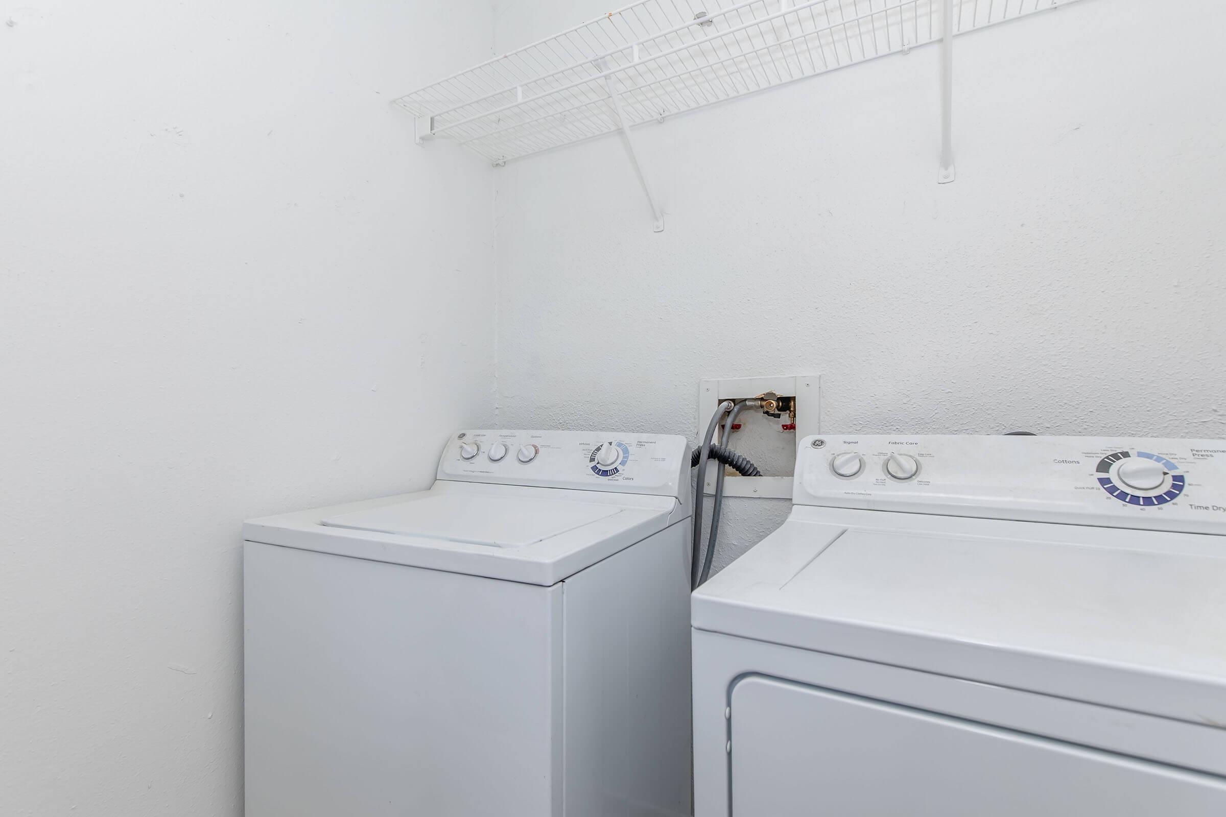A clean, bright laundry room featuring a white washing machine and dryer side by side. Above the appliances, there's a wire shelf for storage. The walls are plain white, creating a simple and functional space for doing laundry.
