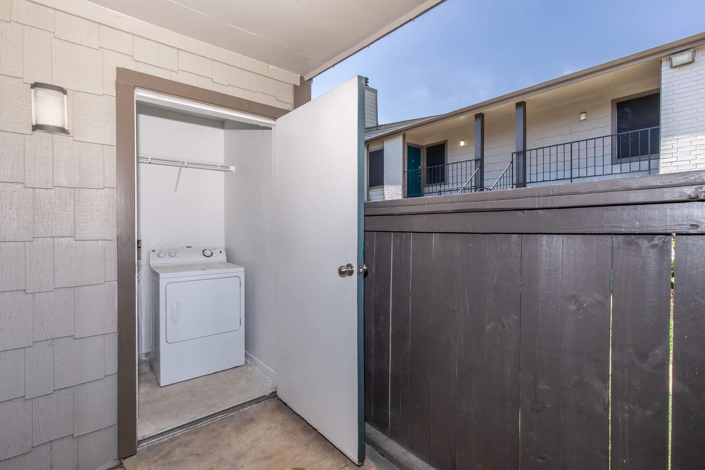 A laundry area with a washer and dryer, visible through an open door, situated next to a wooden fence. In the background, there are apartment buildings with balconies and a clear sky above.