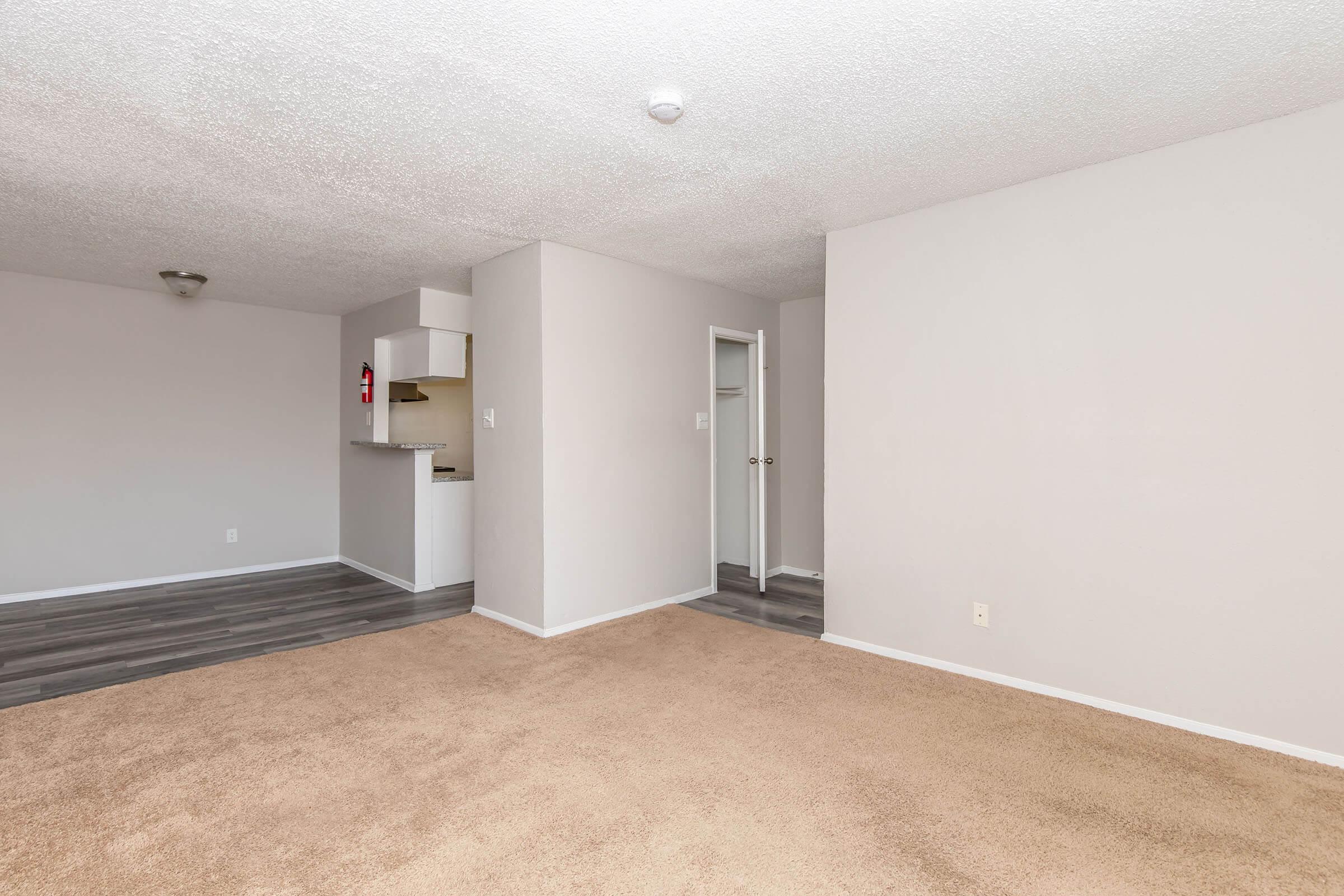 A spacious, empty living room with light beige carpet and neutral-colored walls. In the background, a small kitchenette is visible through an open doorway, featuring a partial wall. The room has a simplified layout with ample natural light and a ceiling light fixture.