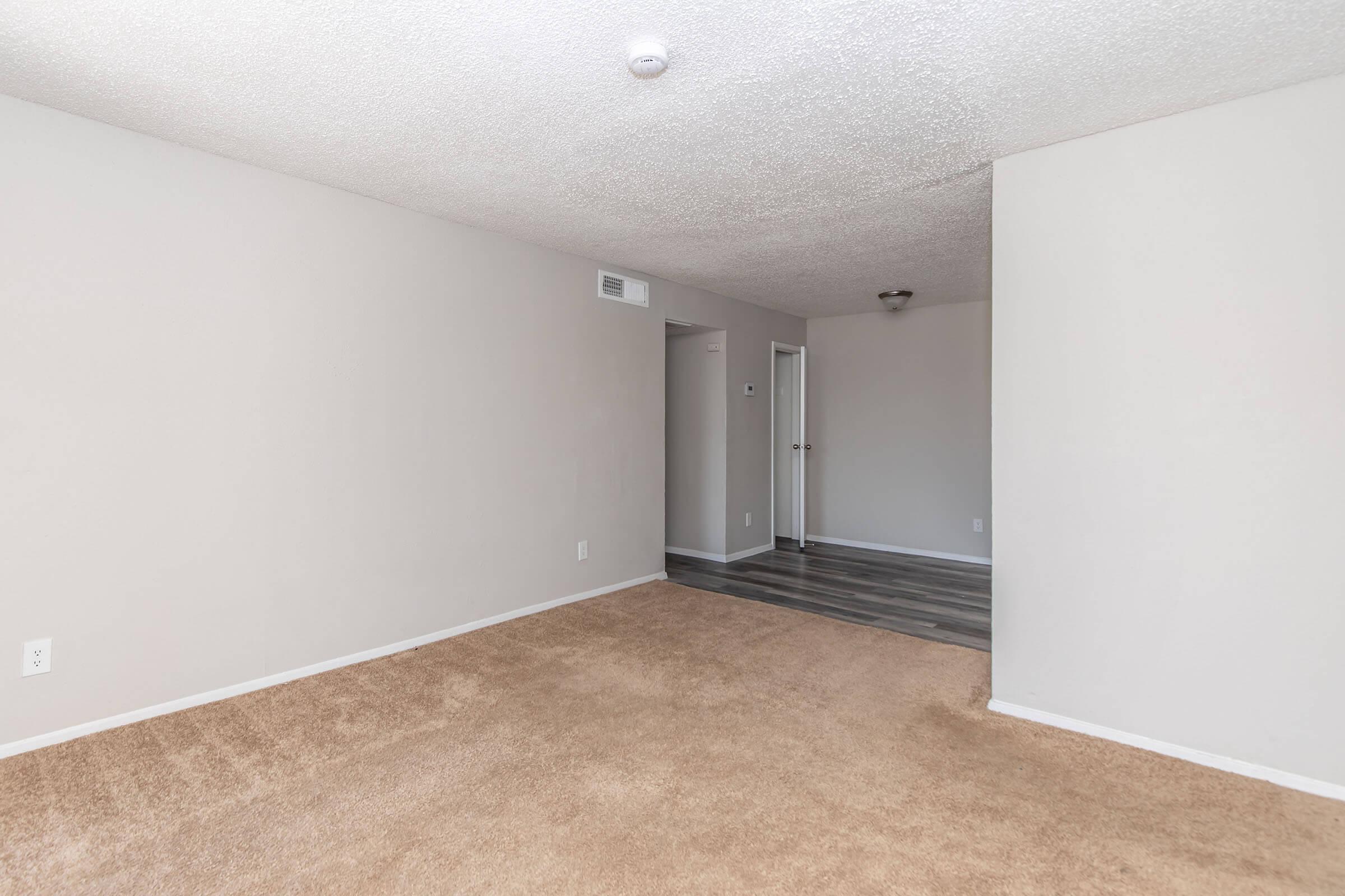 A spacious, empty room with beige carpet and light gray walls. The room features a textured ceiling and a single ceiling light. A doorway leads to a hallway, with another door visible in the background. The room is well-lit and has a clean, minimalistic appearance, ready for decoration.