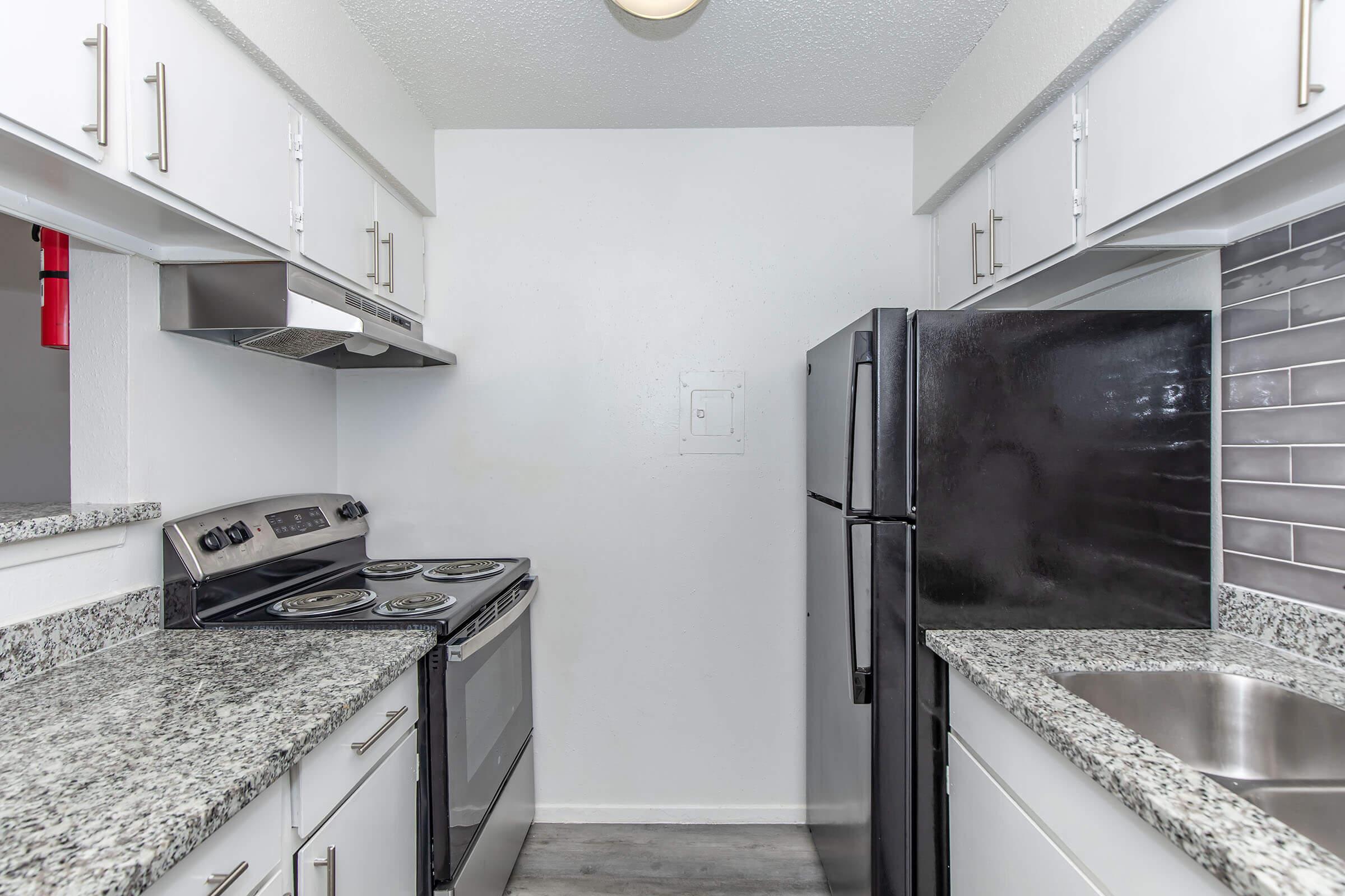 A compact modern kitchen featuring white cabinets, a stainless steel stove and hood, a black refrigerator, and a granite countertop. The walls are painted white, and there is a small sink next to the countertop. The flooring is light-colored, enhancing the bright atmosphere of the space.