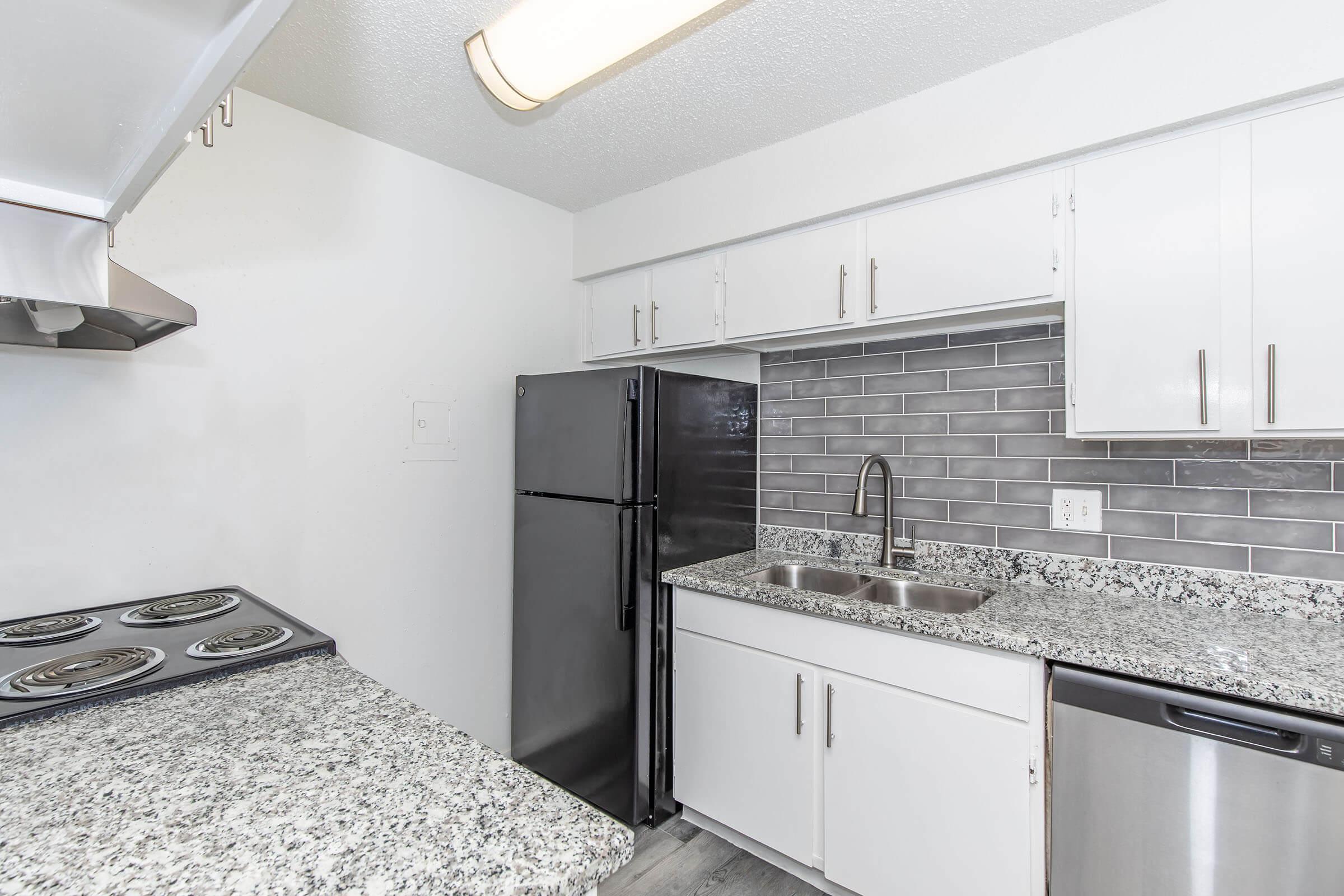 A modern kitchen featuring white cabinetry, a black refrigerator, and stainless steel appliances. The countertops are made of speckled gray granite, and the backsplash consists of gray tiles. Overhead, a simple light fixture illuminates the space. The flooring is light-colored and complements the overall design.