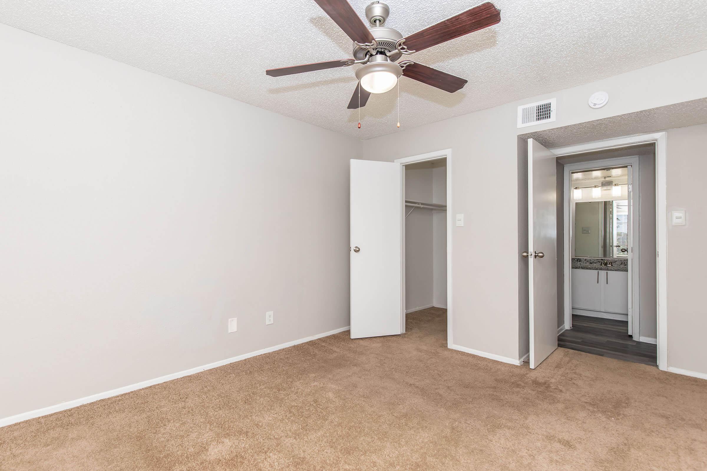 Empty room with light brown carpet, beige walls, and a ceiling fan. One door is open to a closet, while another door leads to an adjoining space. Natural light is visible, and the room appears tidy and well-maintained.