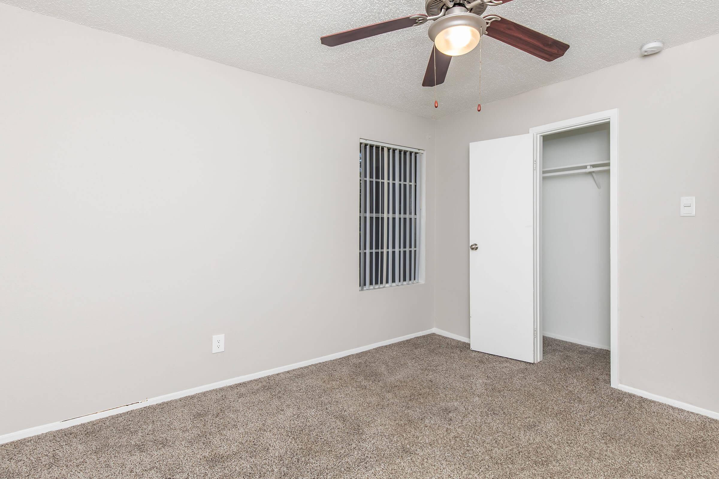 A vacant room with light beige walls and a tan carpet. It features a ceiling fan, a barred window on one side, and a white closet with an open door. The room appears empty, with no furniture or decorations. Overall, it has a clean and simple appearance.