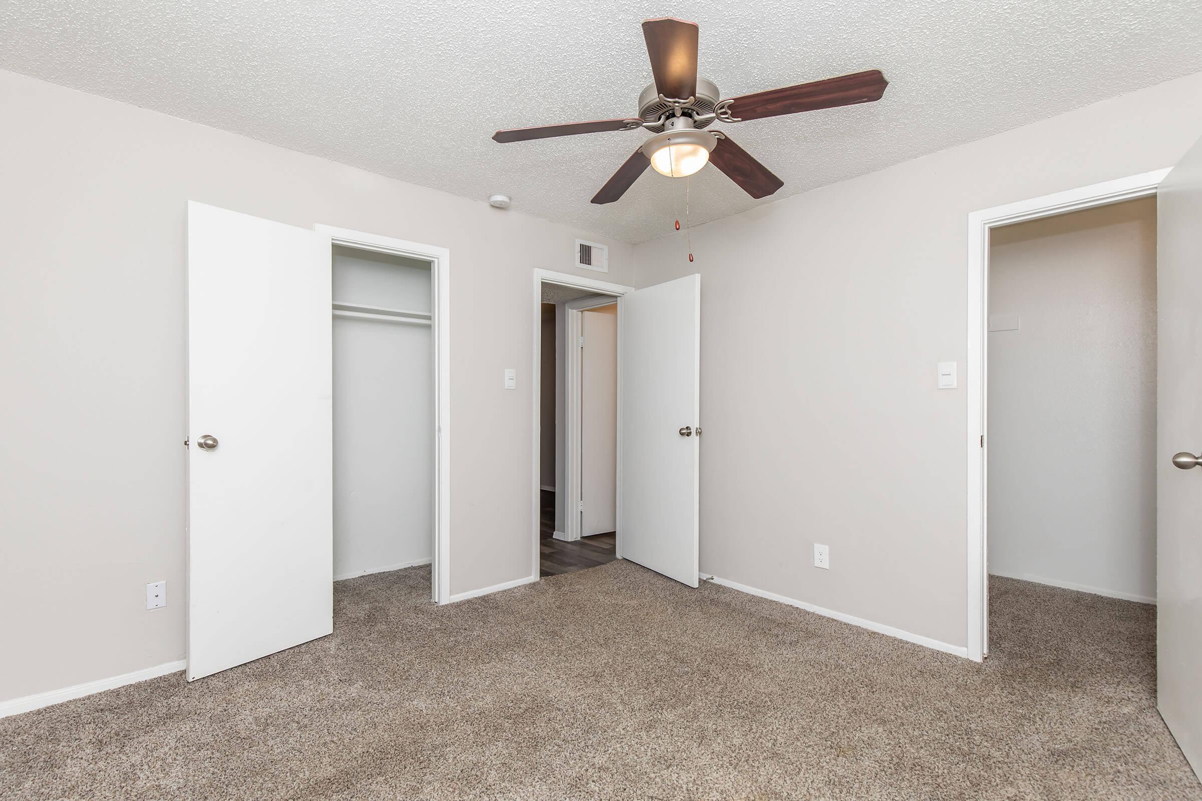Empty room with light beige walls and light brown carpet. A ceiling fan hangs in the center, and there are two white doors leading to closets on opposite walls. The room is well-lit and has a simple, uncluttered aesthetic. The overall appearance is airy and ready for decoration or move-in.