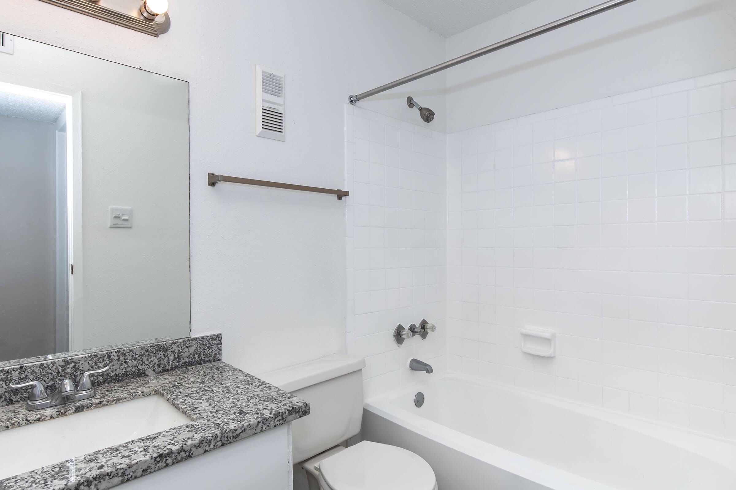 A clean, modern bathroom featuring a white tiled shower/bathtub combination, a granite countertop with a sink, and a wall-mounted mirror. The walls are light-colored, and there is a towel bar beside the bathtub. Natural light enters through a small opening in the adjacent room.