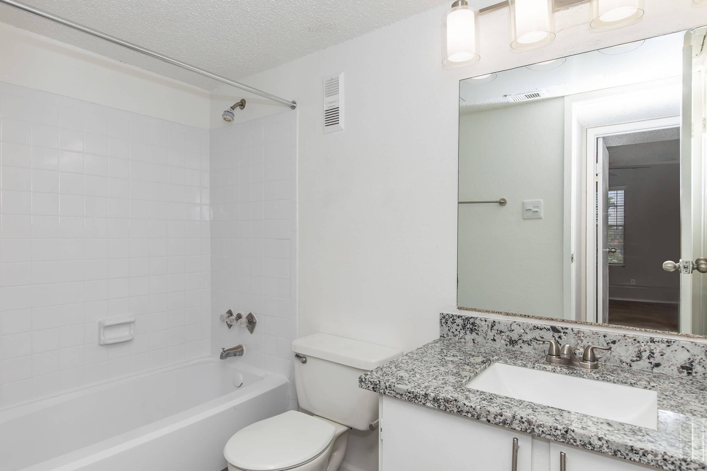 A clean, modern bathroom featuring a white tiled bathtub with a shower, a granite countertop sink with a modern faucet, a toilet, and a well-lit mirror. Light-colored walls and a door leading to another room are visible in the background, creating a spacious feel.