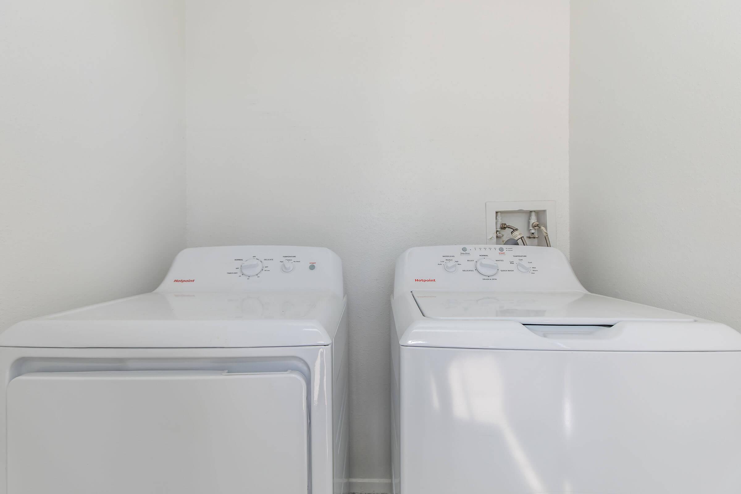 A white washing machine and dryer are positioned side by side against a plain white wall. The machines feature knobs and control panels for settings and operation, highlighting a clean and organized laundry space.