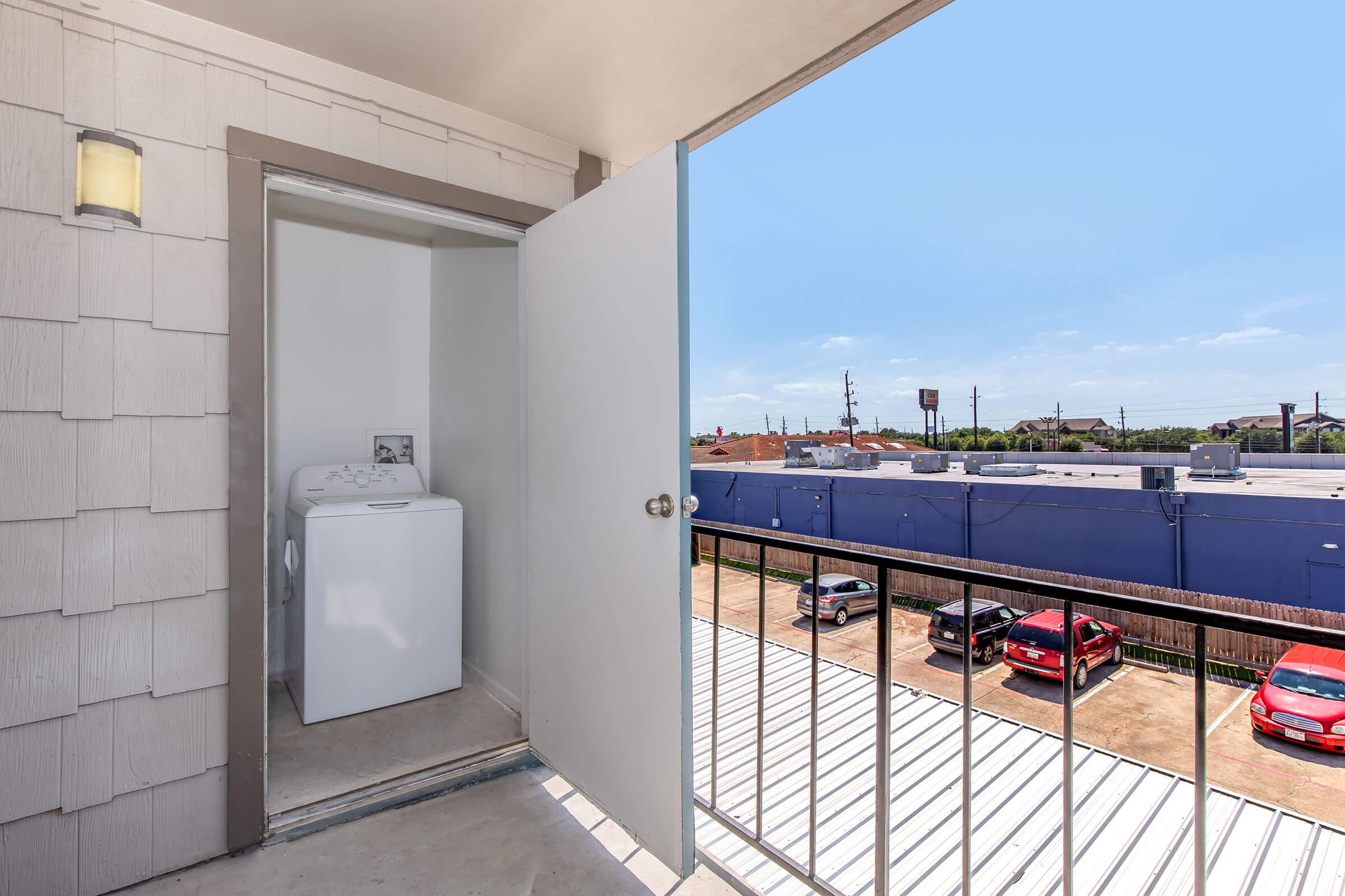 A view from a balcony showing an open door leading to a laundry area with a washing machine. The exterior features a light fixture and a railing. Below, there are parked red cars and a blue building in the background under a clear blue sky.