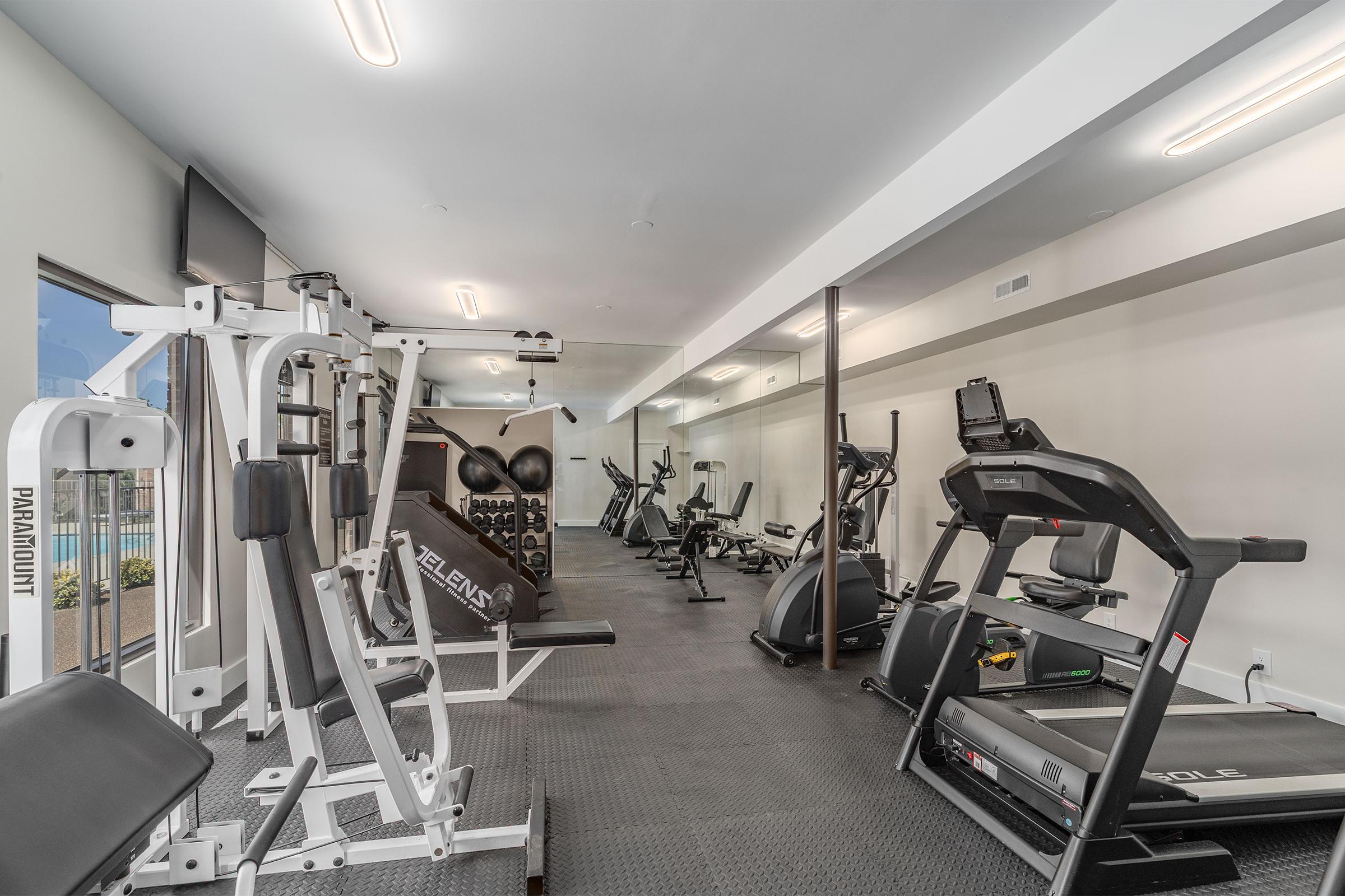 A modern gym interior featuring various fitness equipment, including weightlifting machines, treadmills, and elliptical machines. The space is well-lit with overhead lights and includes mirrors along the walls. There is a clean, organized layout with rubber flooring.
