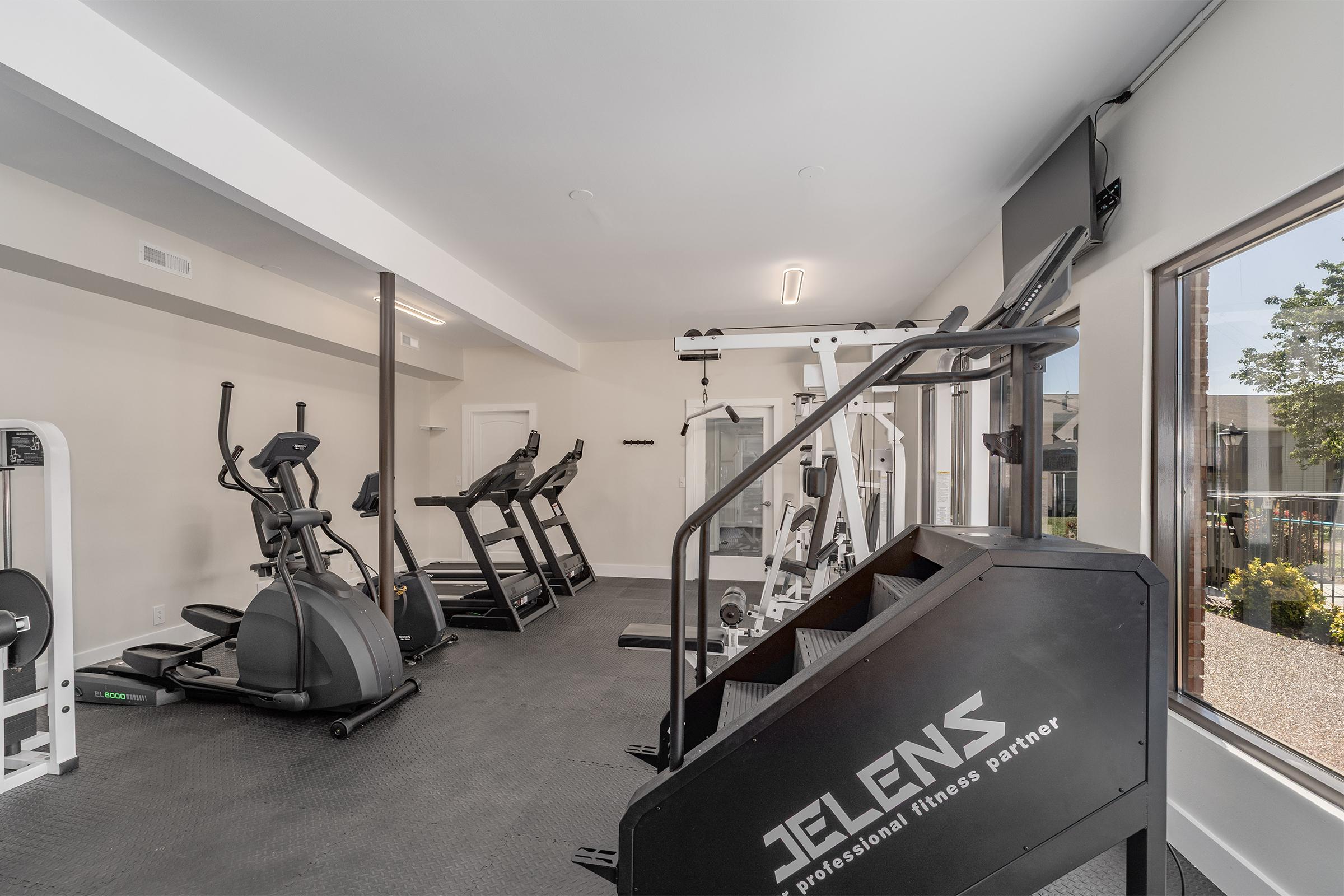 A modern gym interior featuring various fitness equipment, including treadmills, an elliptical machine, a strength training machine, and a stair climber. The space has a sleek design with large windows allowing natural light, enhancing the workout atmosphere. Flooring is black rubber matting for safety and comfort.