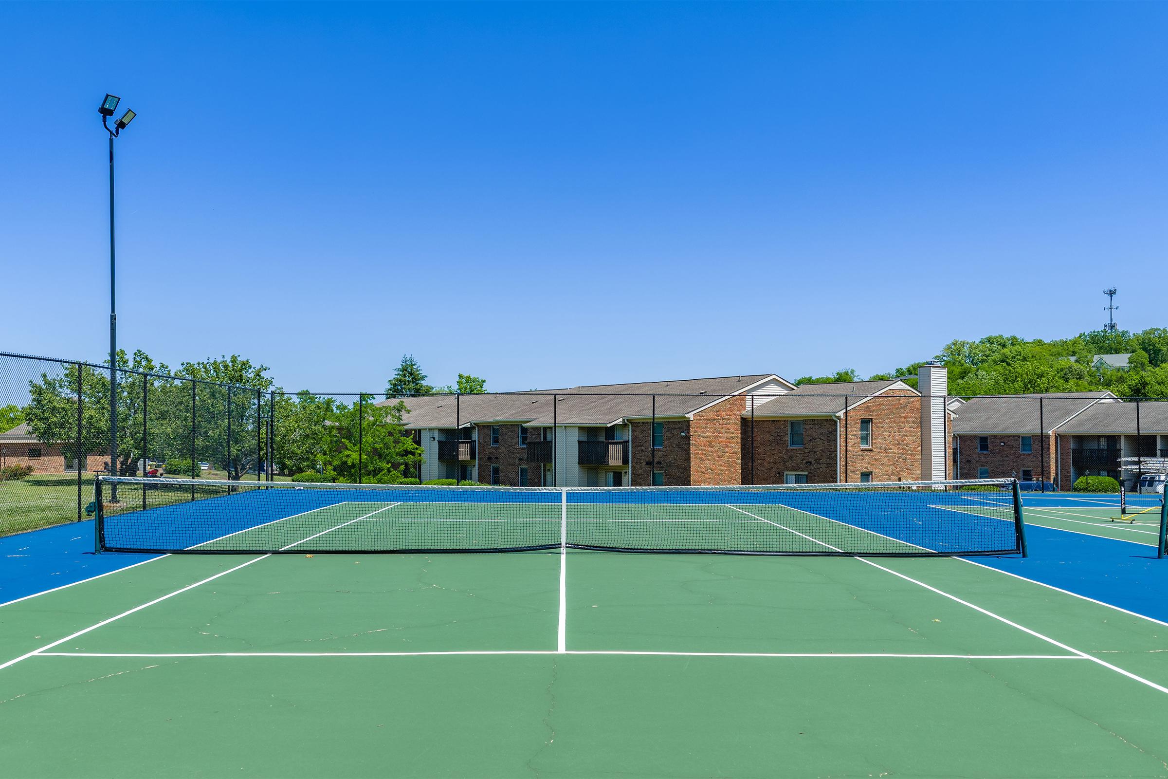 A bright, clear day at a tennis court with a green playing surface and a net in the foreground. In the background, there are several brick apartment buildings surrounded by a grassy area and trees. The sky is clear blue, and there are sports lights on poles surrounding the court.