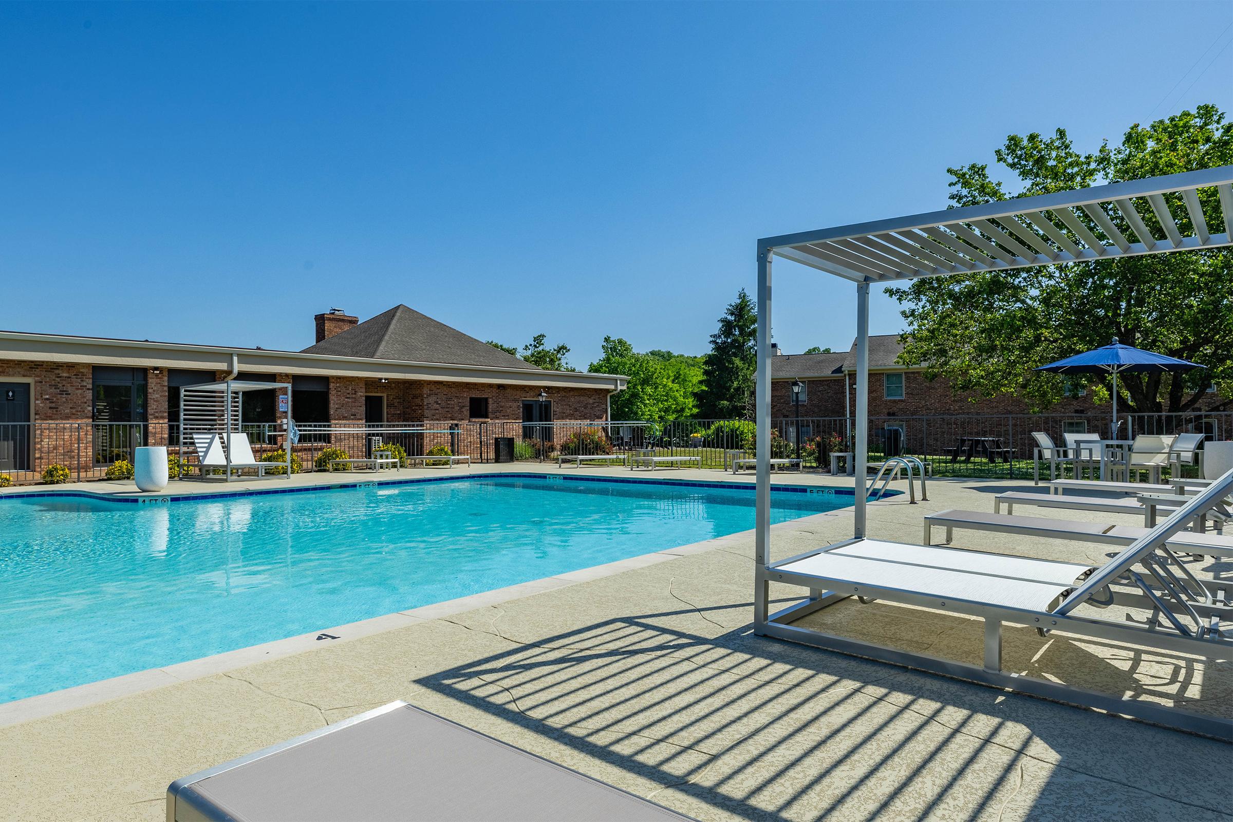 A clear blue swimming pool surrounded by lounge chairs and a pergola, with apartment buildings in the background under a bright blue sky. Trees provide shade near the pool area, creating a relaxing outdoor space for residents.