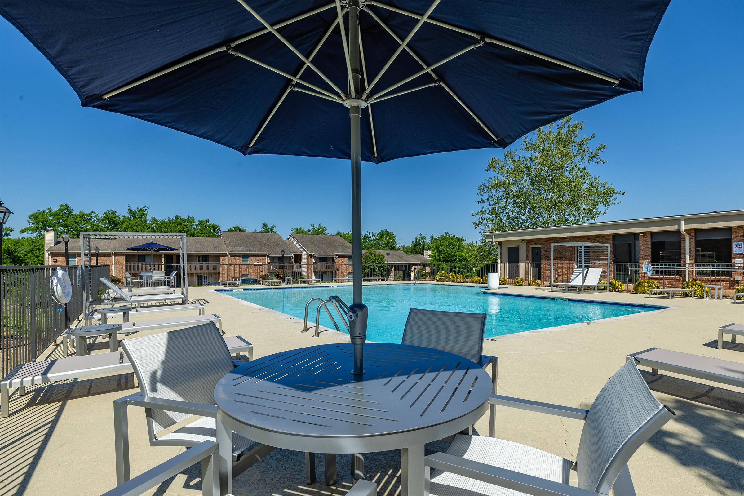 A sunny outdoor pool area featuring a large swimming pool, surrounded by lounge chairs and tables with umbrellas. In the background, there are residential buildings and lush green trees. The clear blue sky enhances the inviting atmosphere of the space.