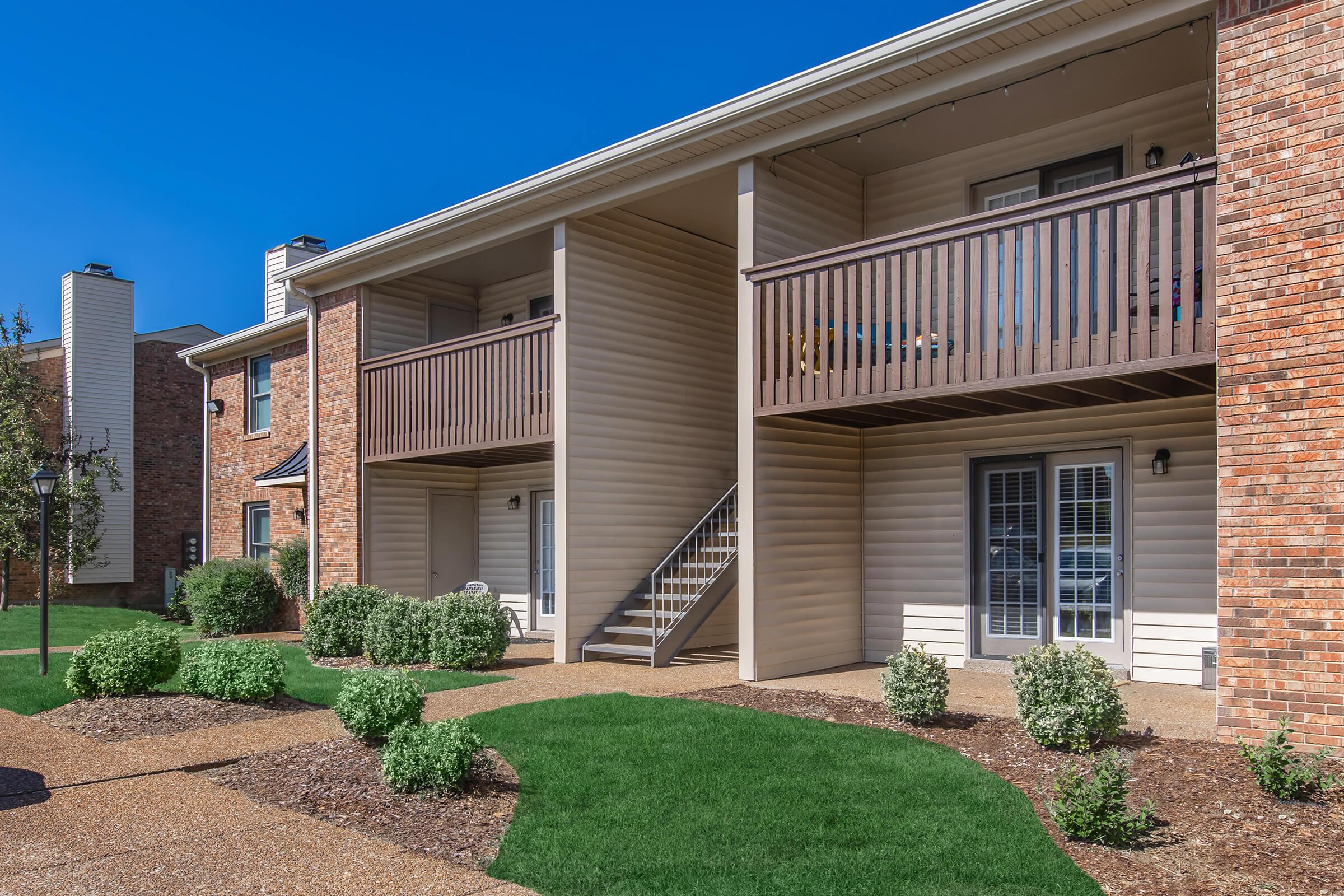 A well-maintained apartment complex featuring two-story buildings. The exterior includes wooden balconies and stairs, surrounded by neatly trimmed grass and small shrubs. The sky is clear and blue, creating a bright, inviting atmosphere.